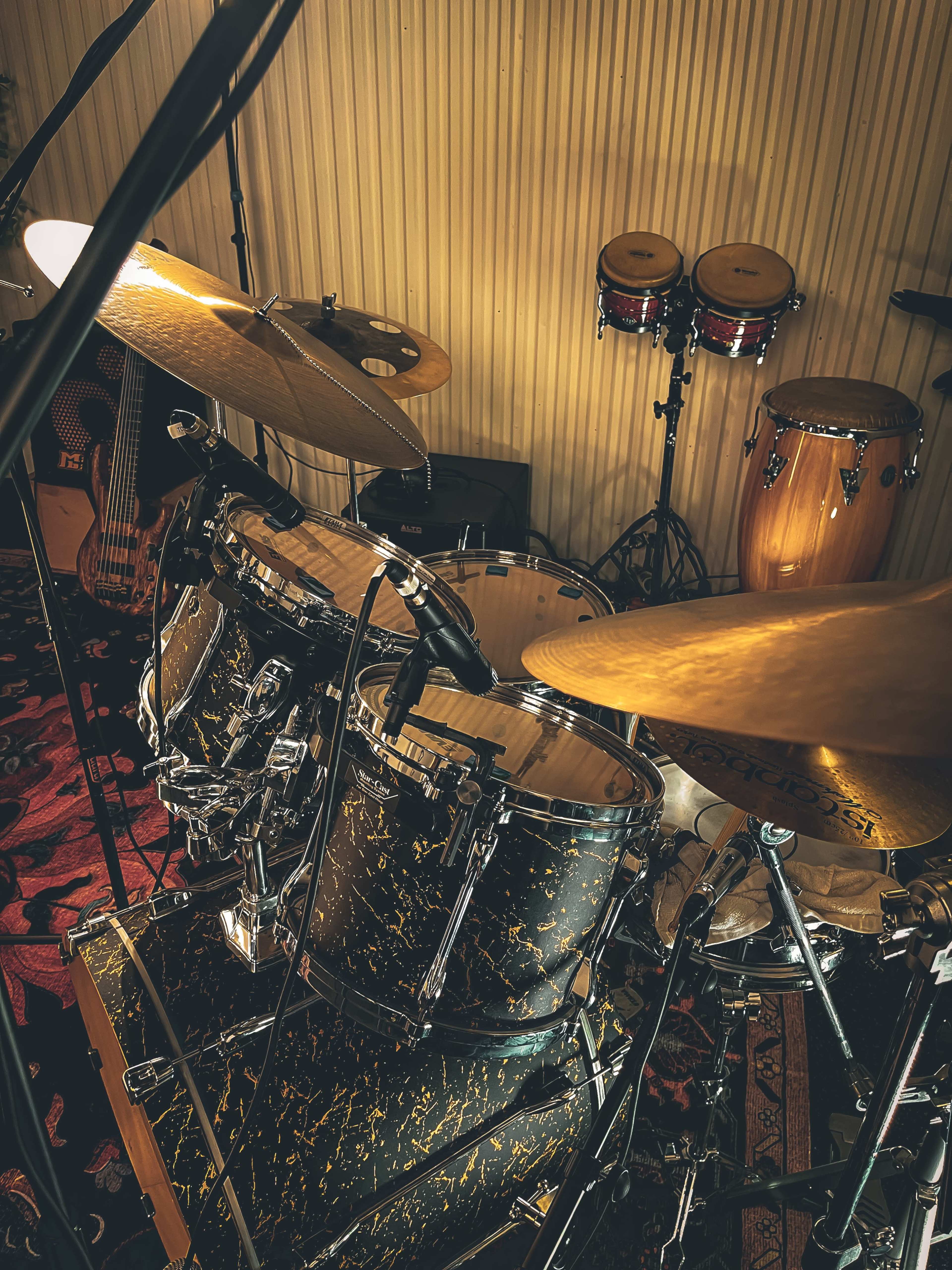 The image shows a close-up view of a drum set with cymbals, surrounded by various percussion instruments in a recording studio setting.