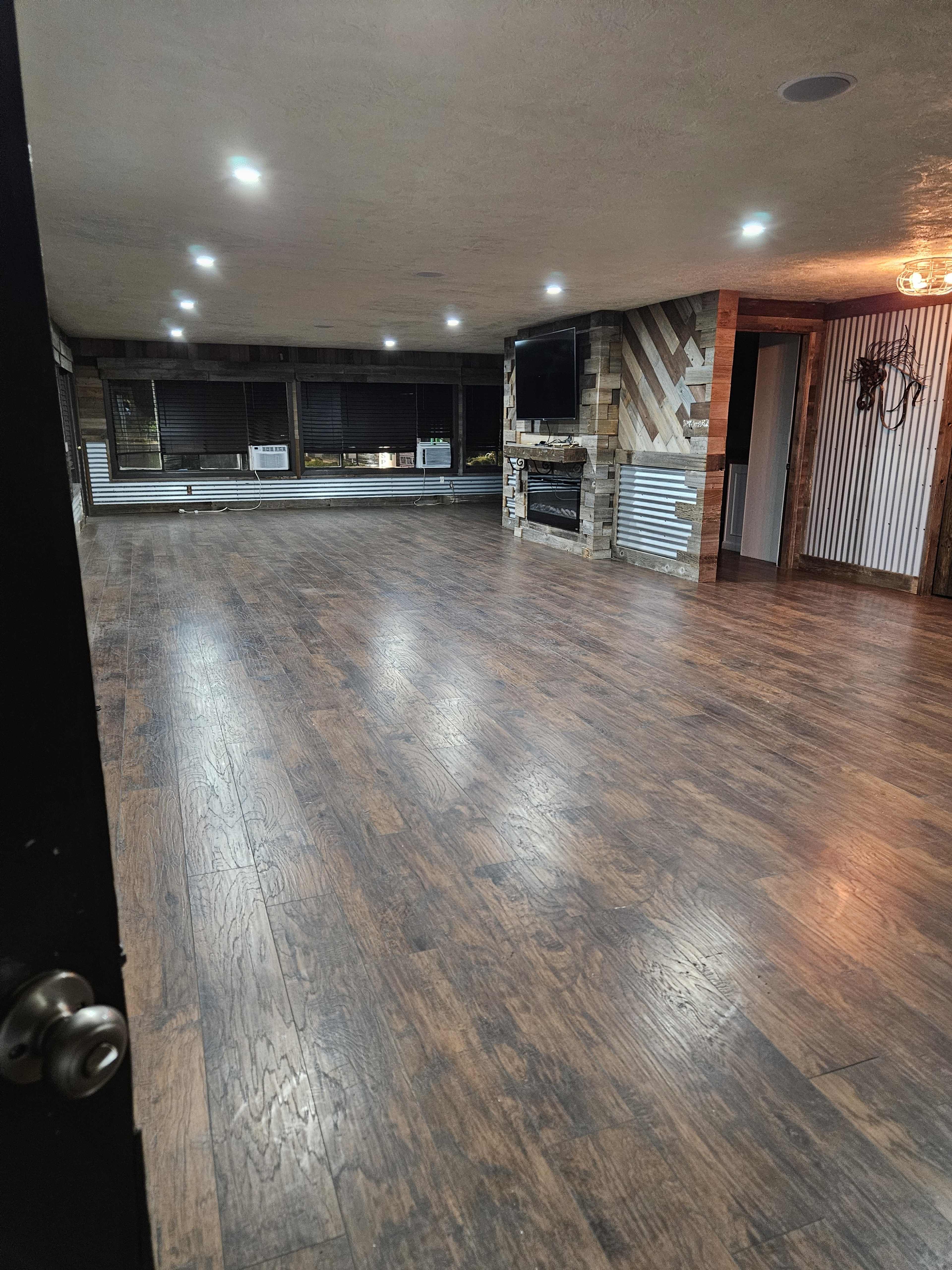 The image shows a spacious room with wooden flooring, modern lighting, and a brick fireplace.