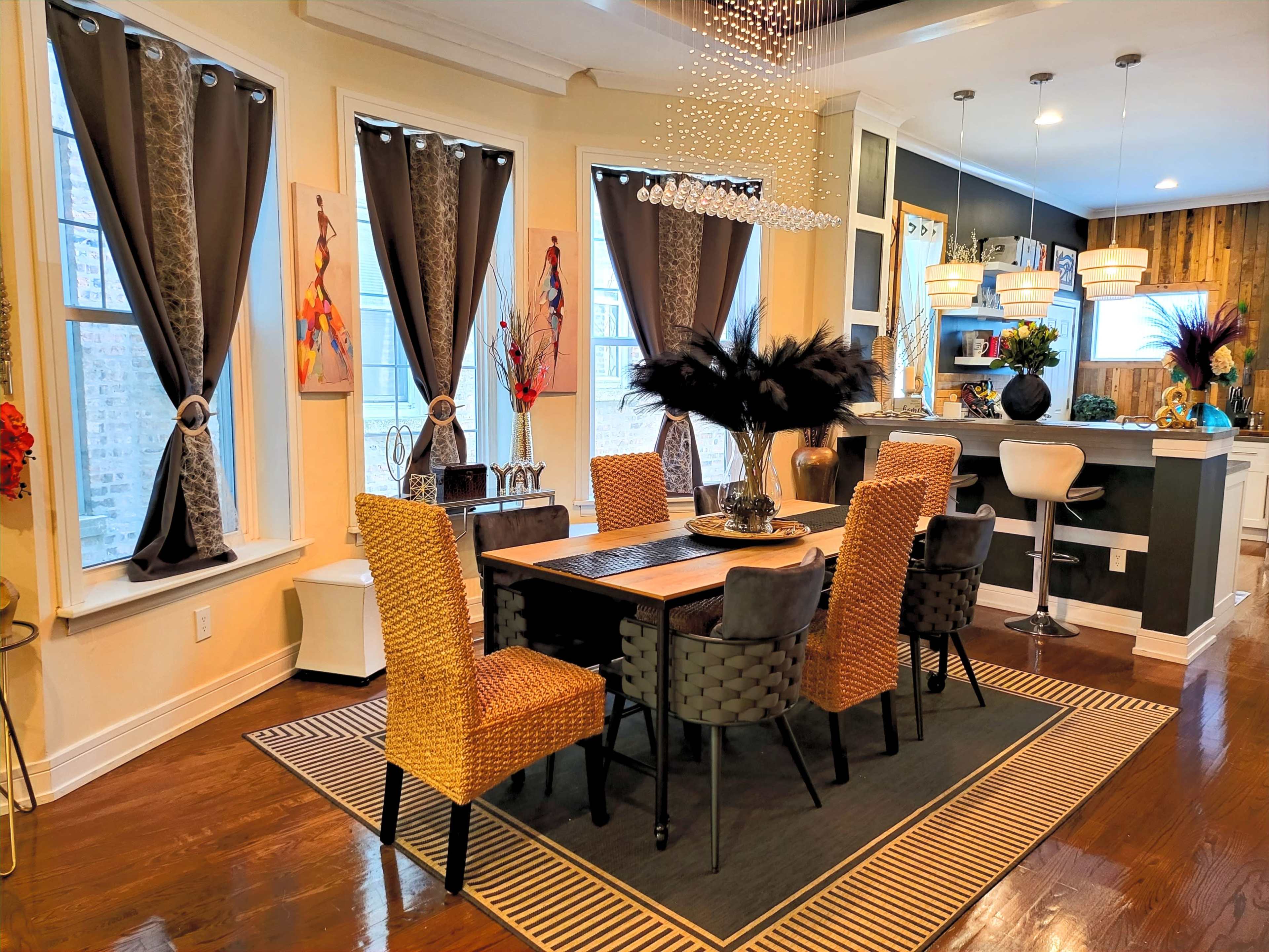 A dining area features a wooden table surrounded by woven and upholstered chairs, with large windows draped in dark curtains and a modern kitchen visible in the background.