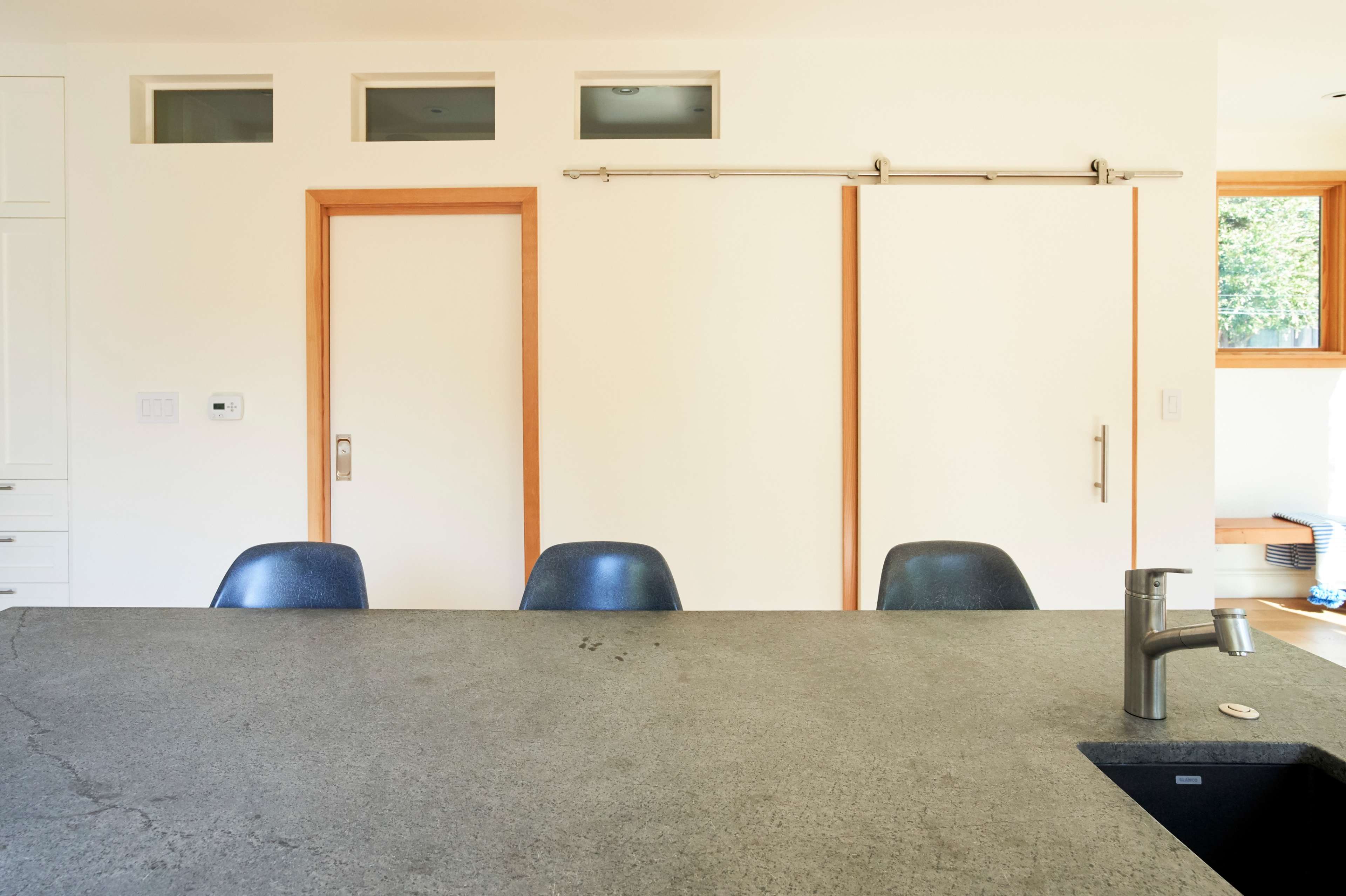A minimalist kitchen with a concrete countertop, three blue chairs in front of it, and sliding doors on both sides of a central door.
