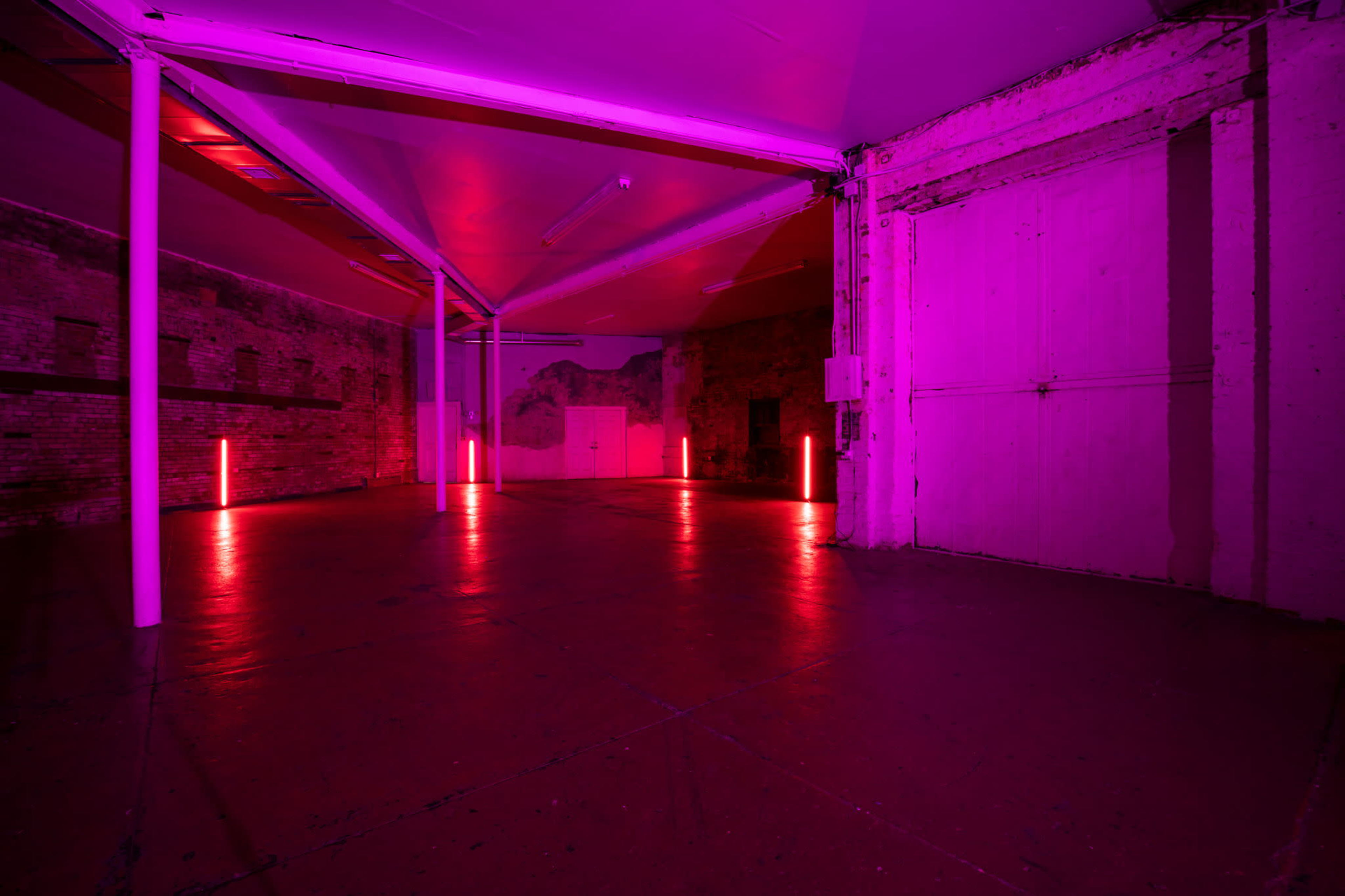 The image shows an empty industrial space illuminated with pink lighting, featuring exposed brick walls and a concrete floor.