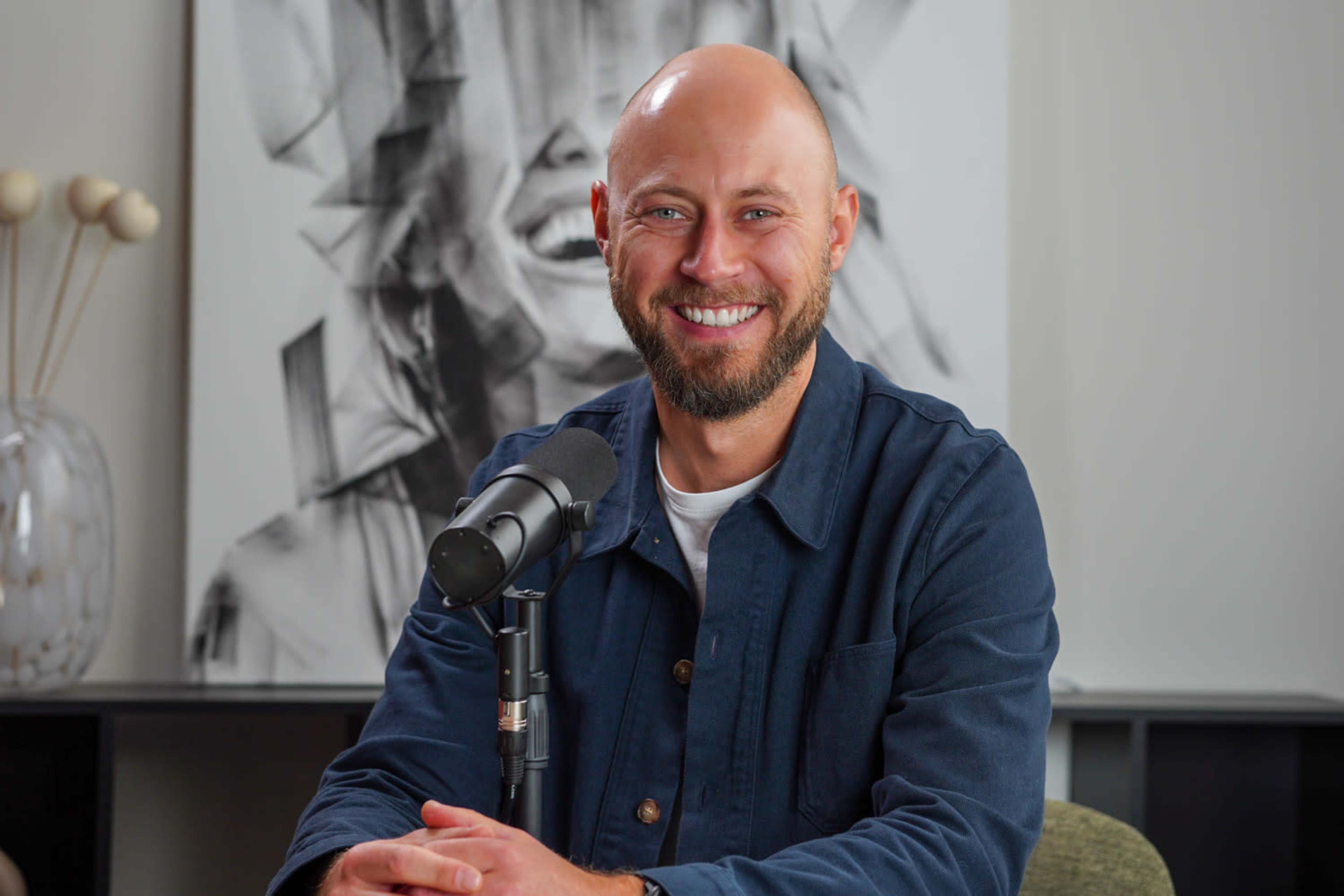 A man with a bald head and beard smiles while sitting in front of a microphone and a large artistic portrait in a well-lit room.