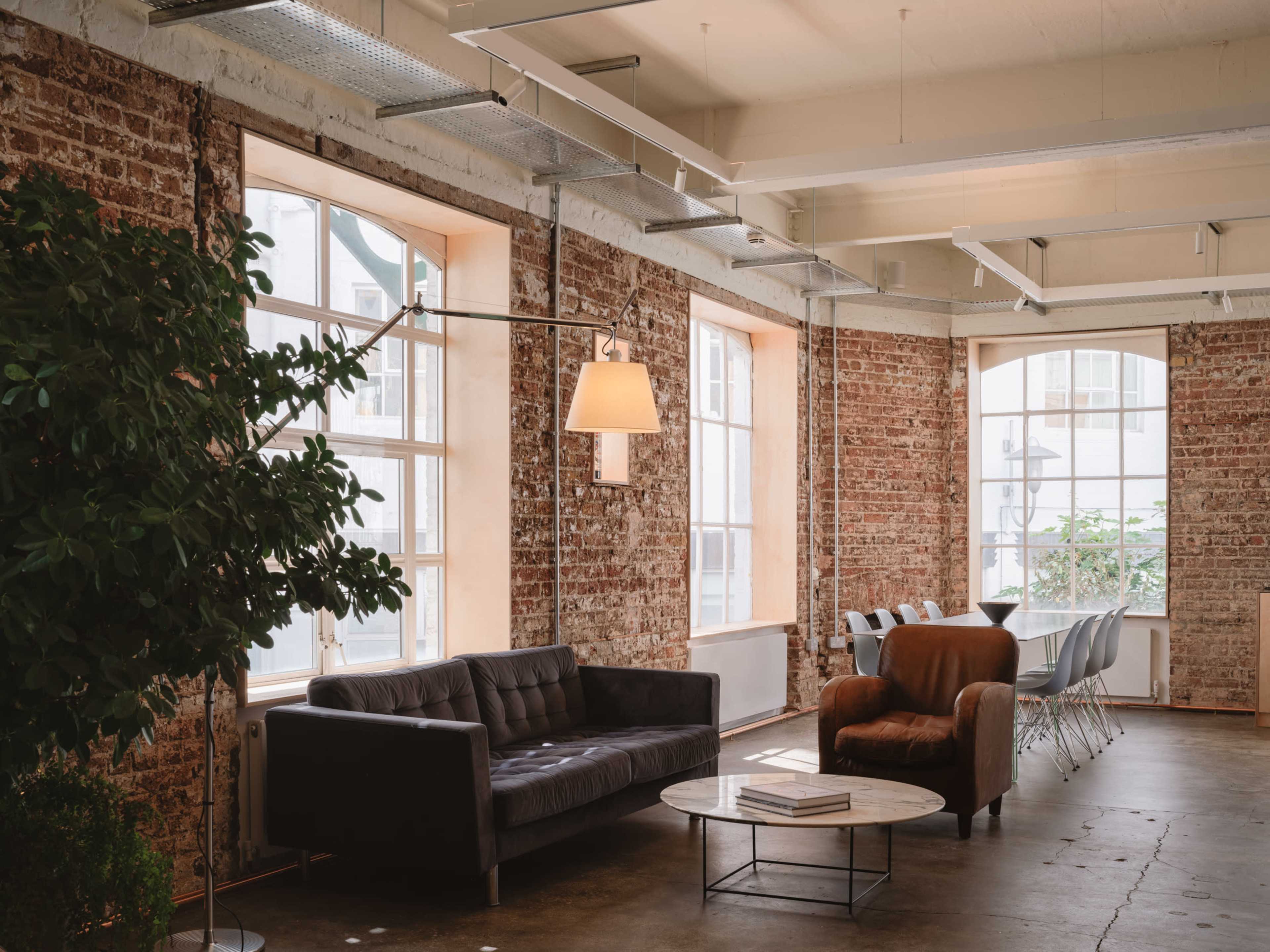 The image shows a modern workspace with exposed brick walls, a leather couch, an armchair, a coffee table, and large windows allowing natural light to fill the room.