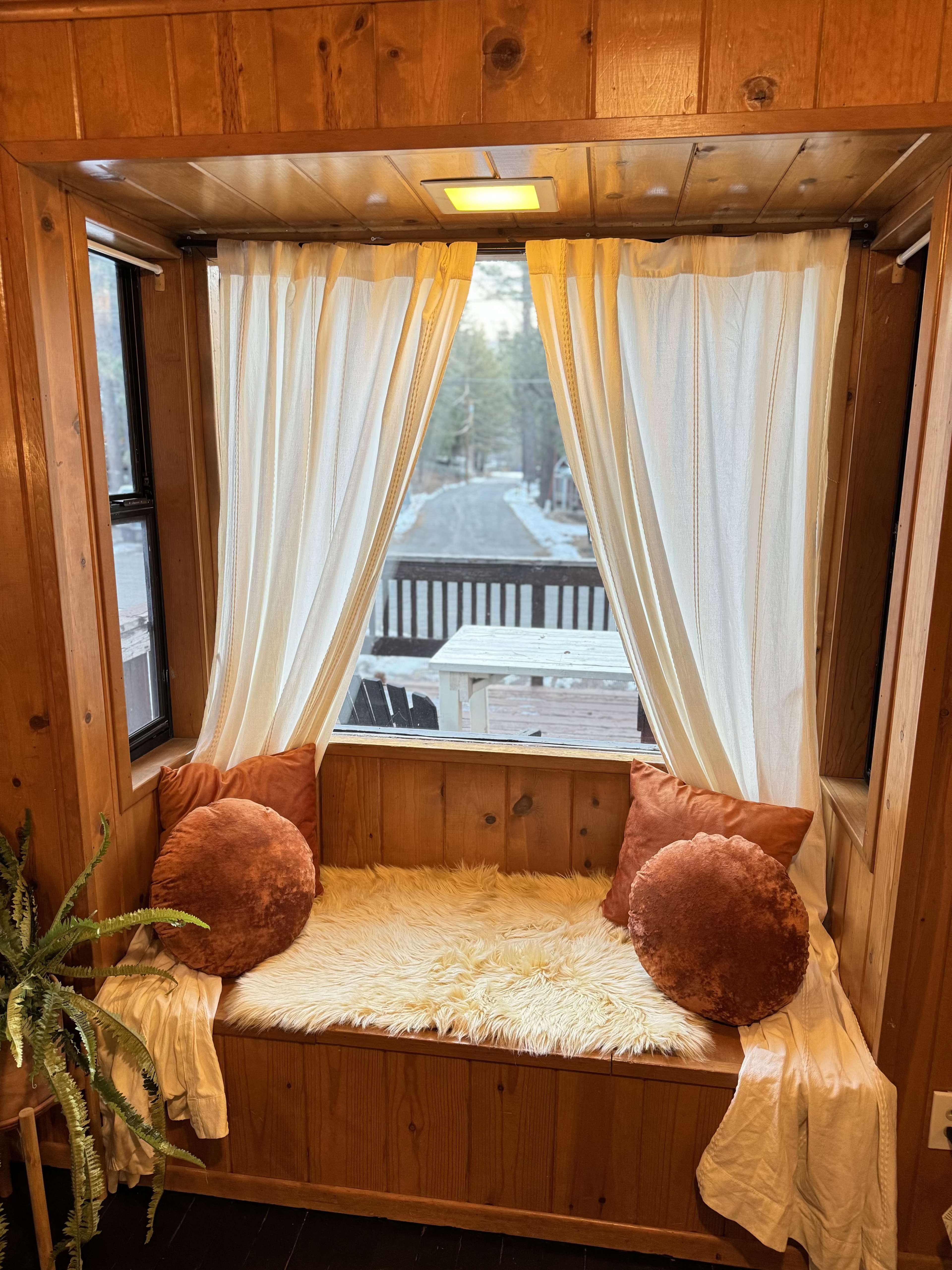 A cozy window nook features soft pillows and a fluffy throw, framed by light curtains and wooden walls, with a view of an outdoor deck.