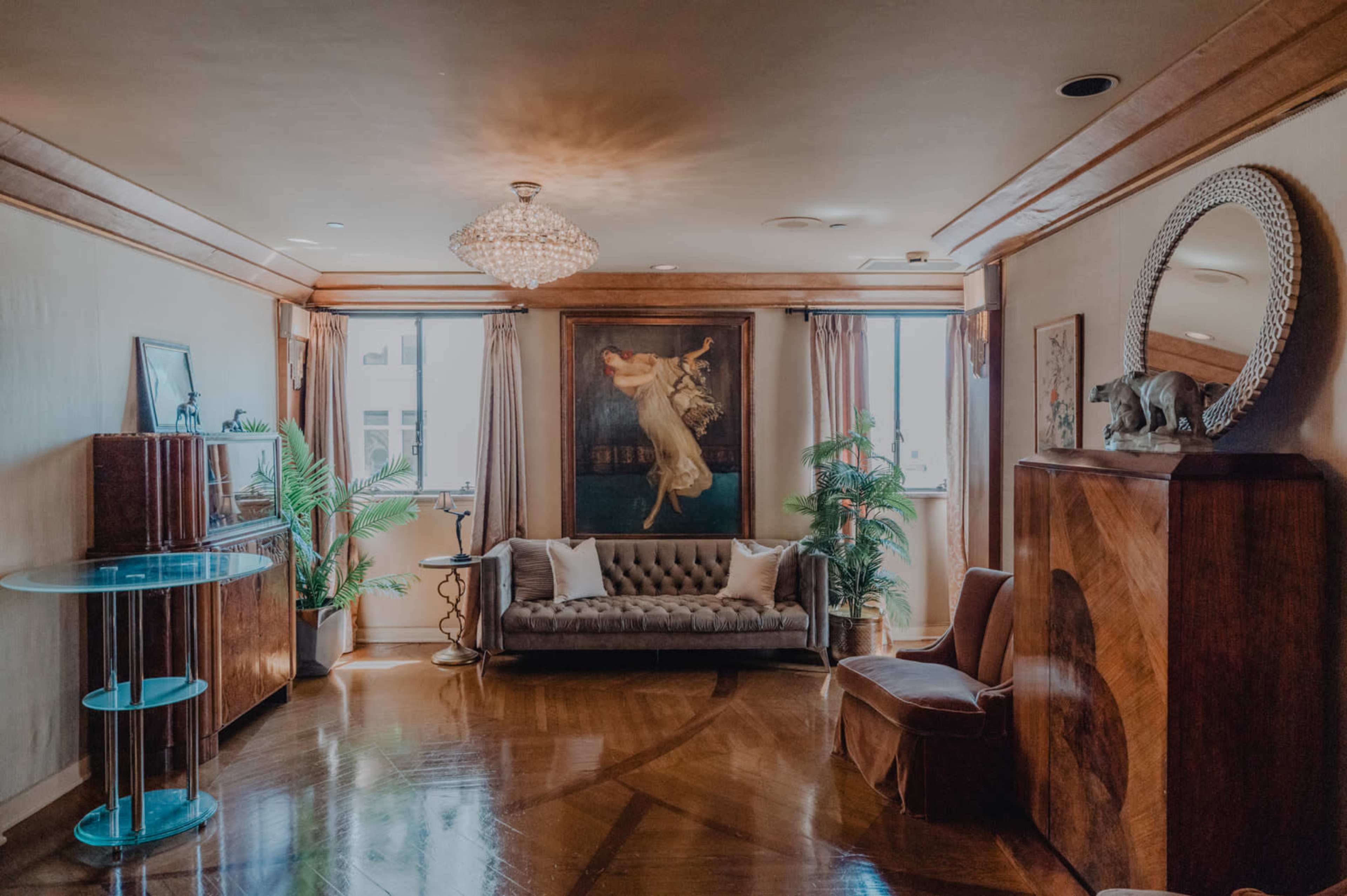 The image shows a stylish living room featuring a plush sofa, a large artwork on the wall, and decorative elements including plants and a chandelier.