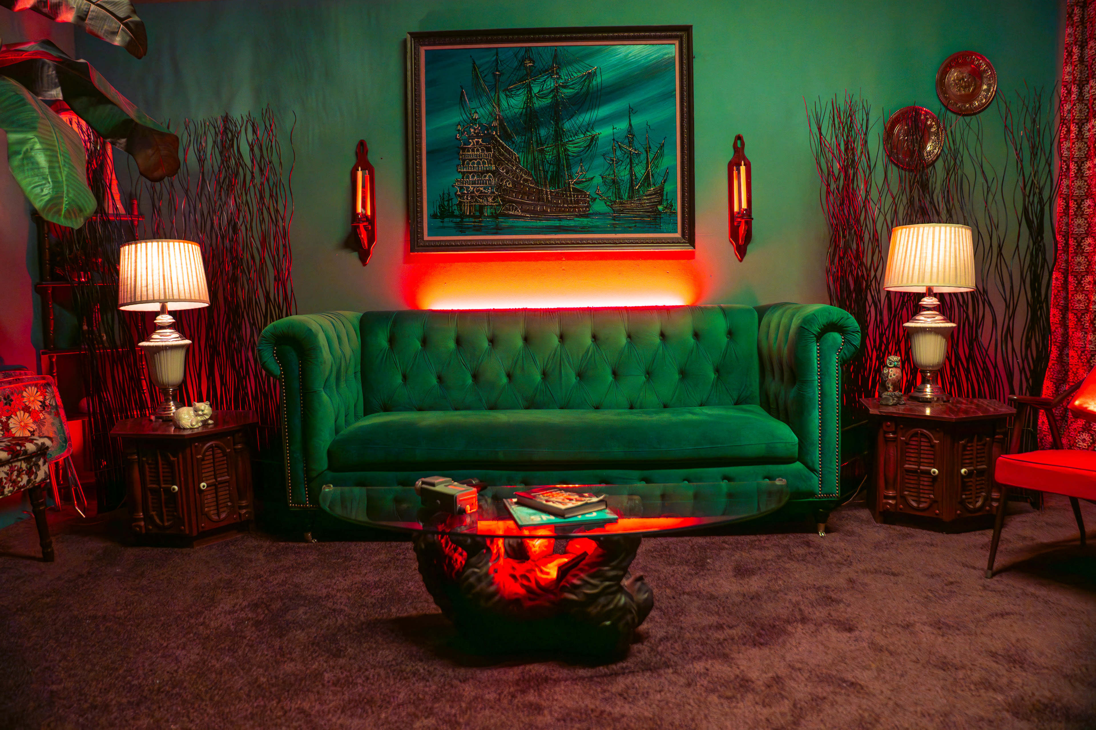 The image shows a vintage-style living room featuring a green tufted sofa, a glass coffee table, decorative lamps, and a painting of a ship on the wall, all set against a dimly lit backdrop with plants and red accents.