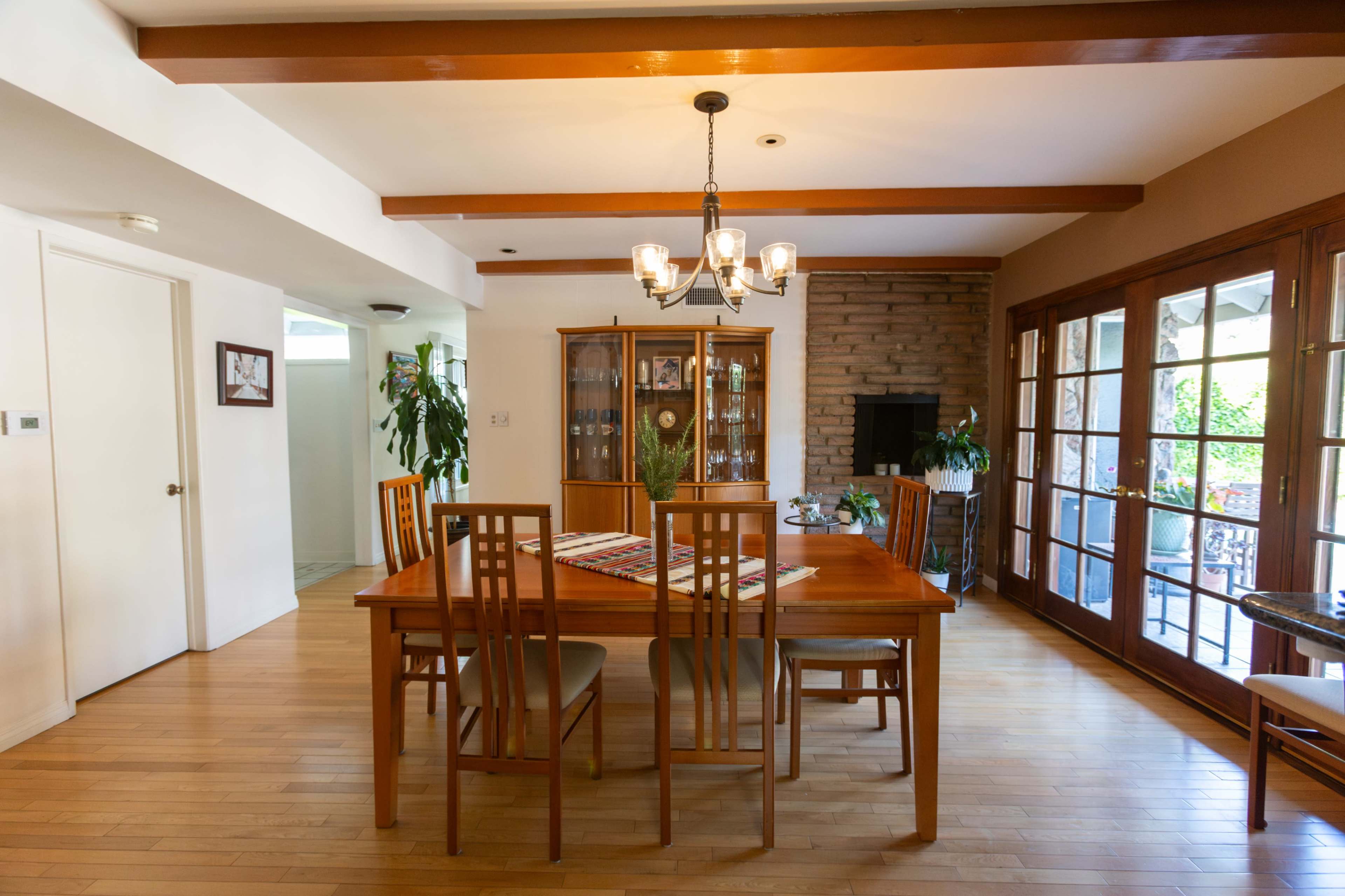 A dining room features a wooden table surrounded by chairs, with a display cabinet and large windows leading to an outdoor patio.