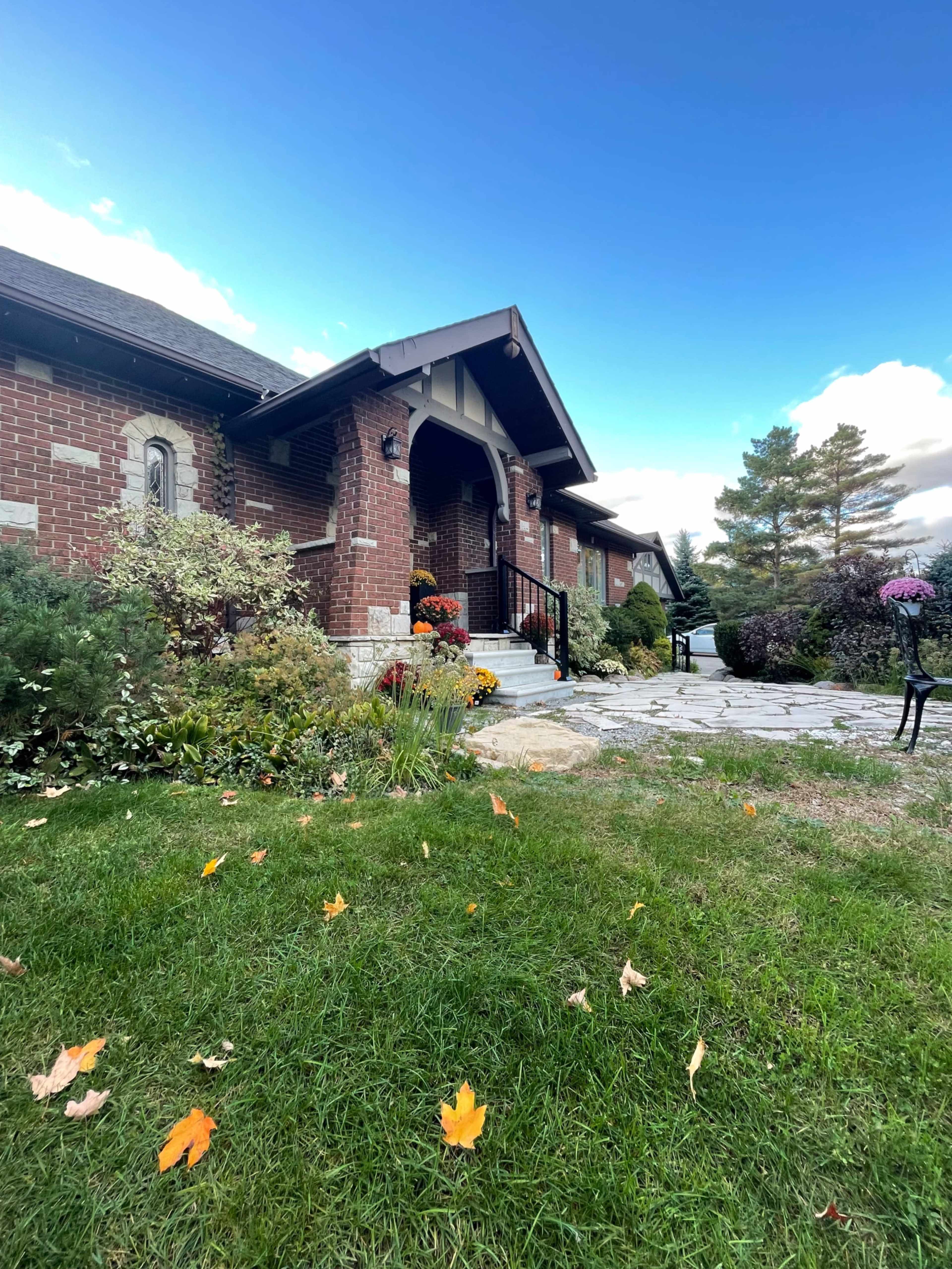 A brick house with a front porch is surrounded by greenery and autumn leaves on the lawn.