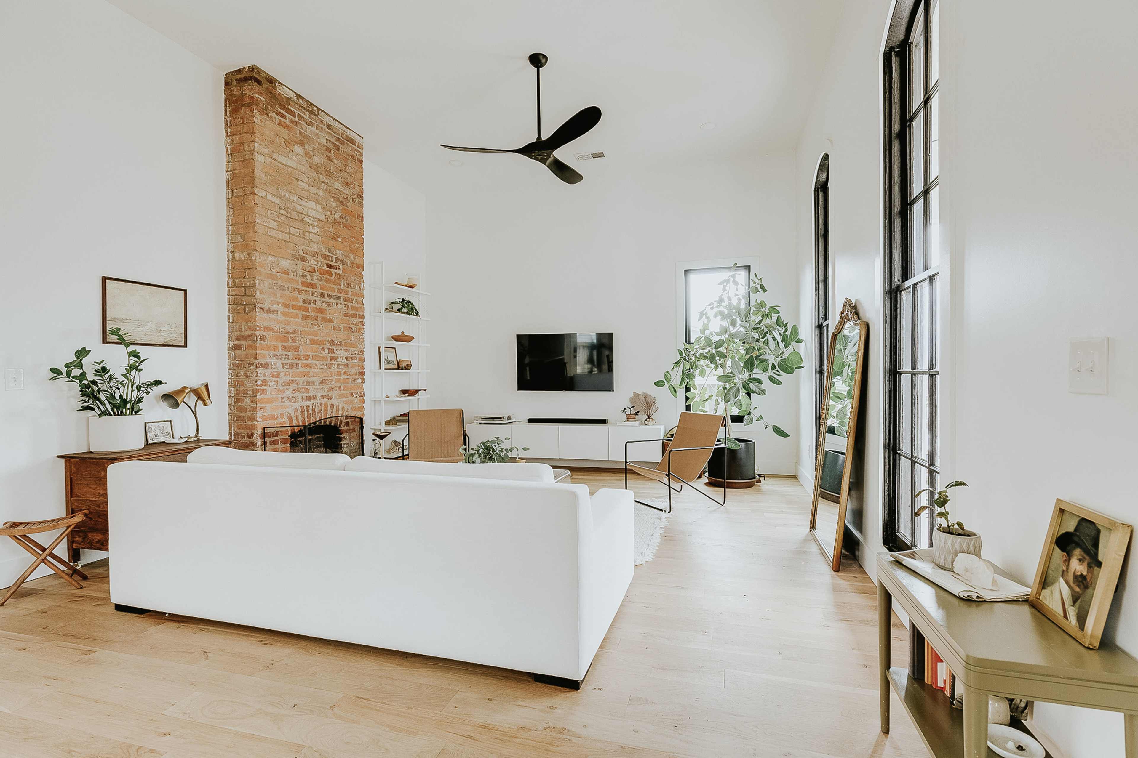 A modern living room features a white sofa, a brick accent wall, a flat-screen TV, and large windows with plants.