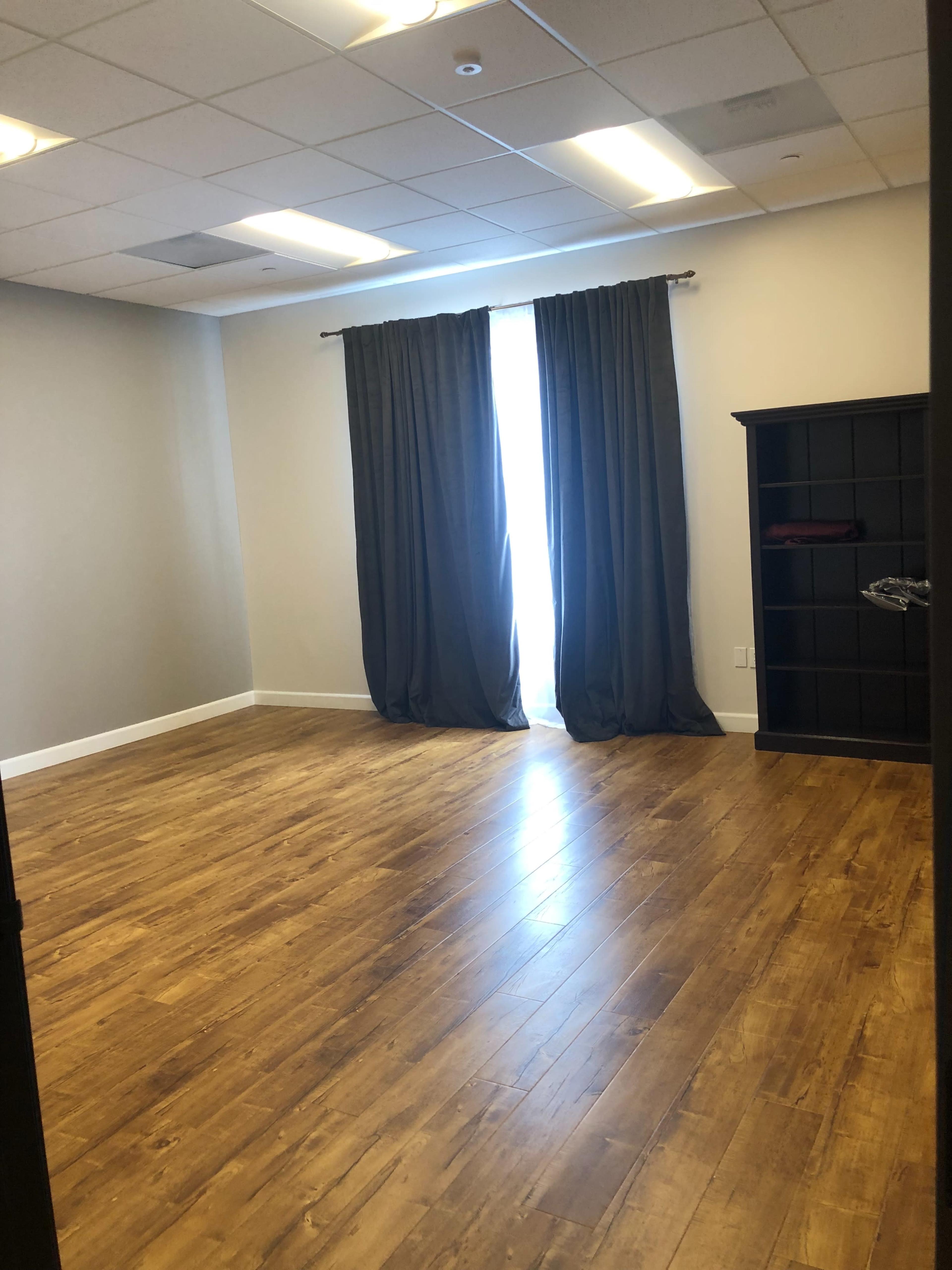 The image shows a sparsely furnished room with wooden flooring, a large window covered by dark curtains, and a black shelving unit against one wall.
