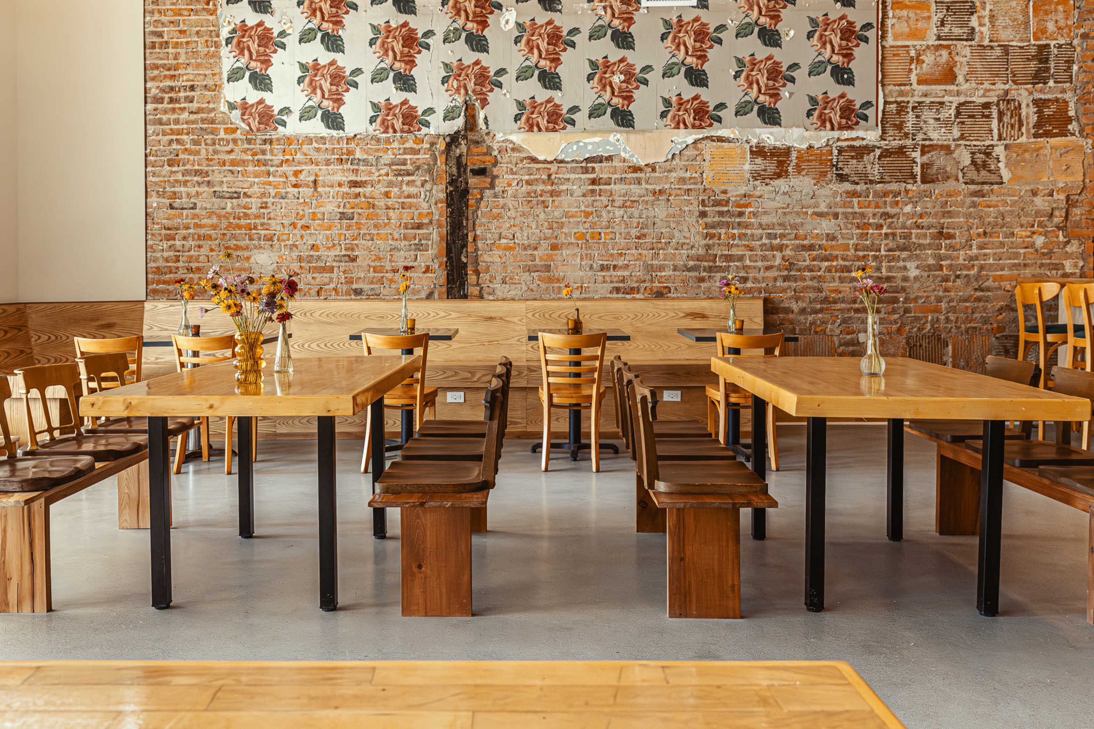 A bright interior of a restaurant features wooden tables and chairs arranged on a polished floor against a backdrop of exposed brick and floral wallpaper.