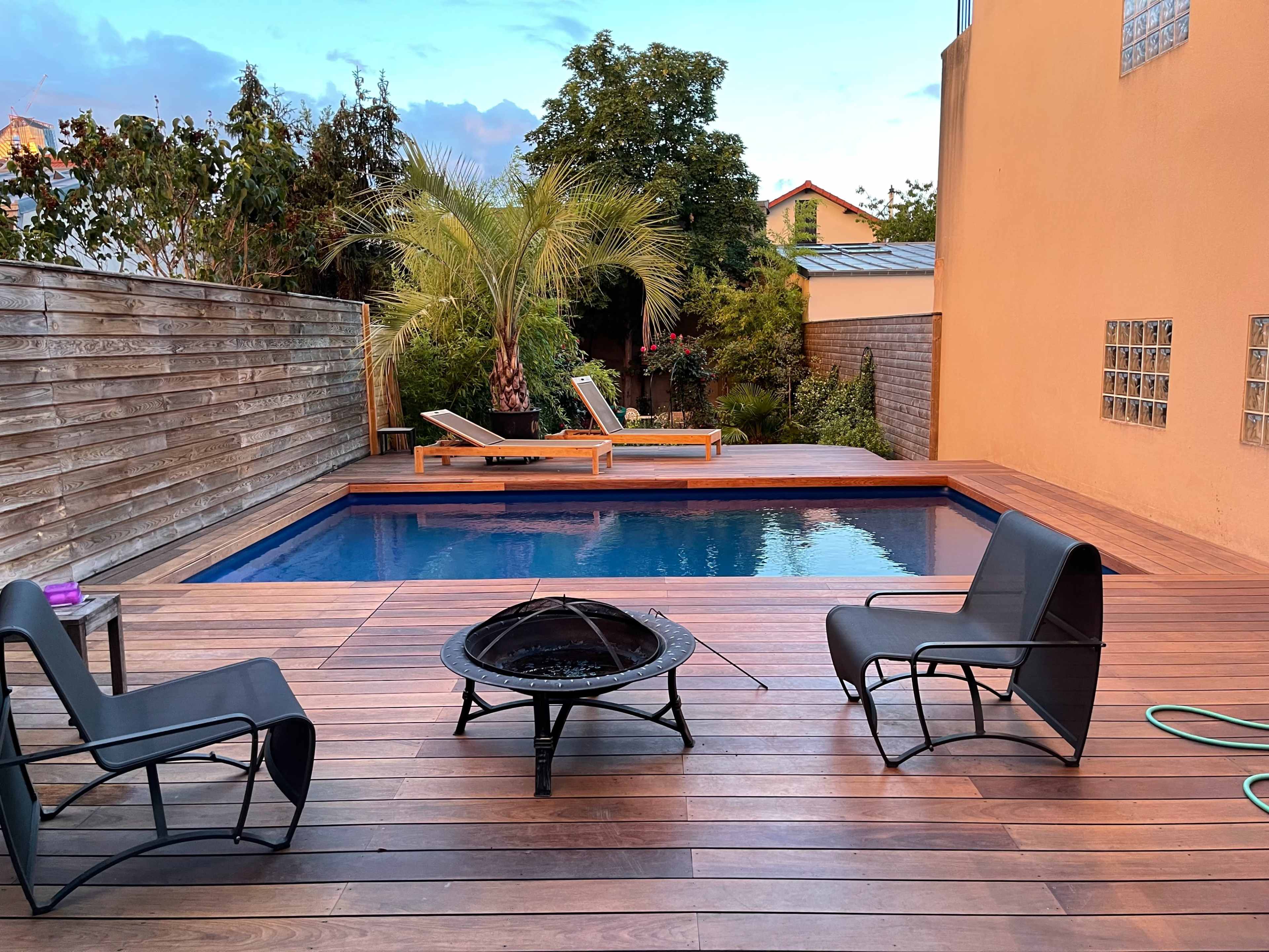 A backyard area featuring a small pool surrounded by wooden decking, two lounge chairs, and a fire pit.