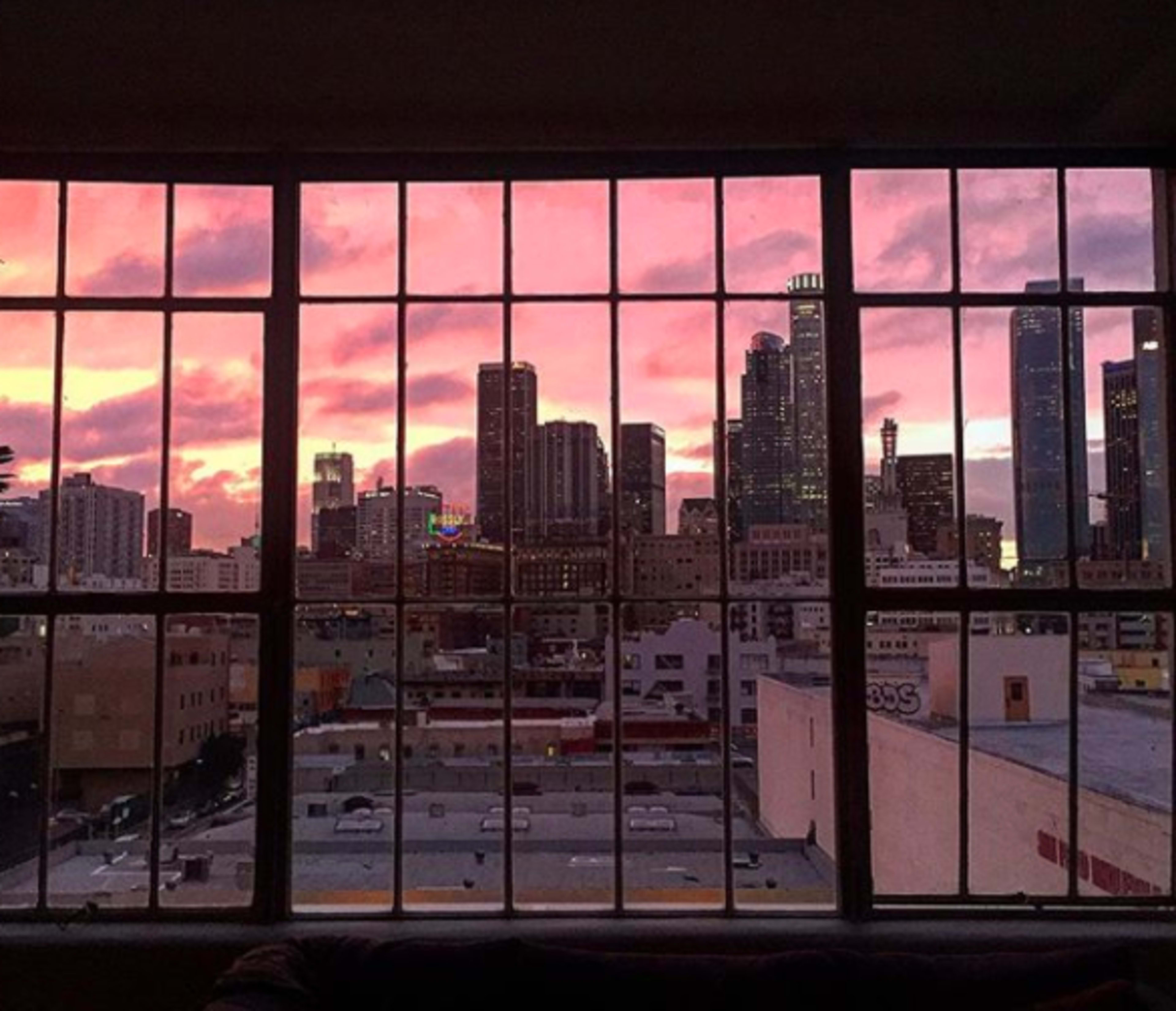 A city skyline is framed by a large window, with a vibrant sunset casting pink and orange hues across the buildings.