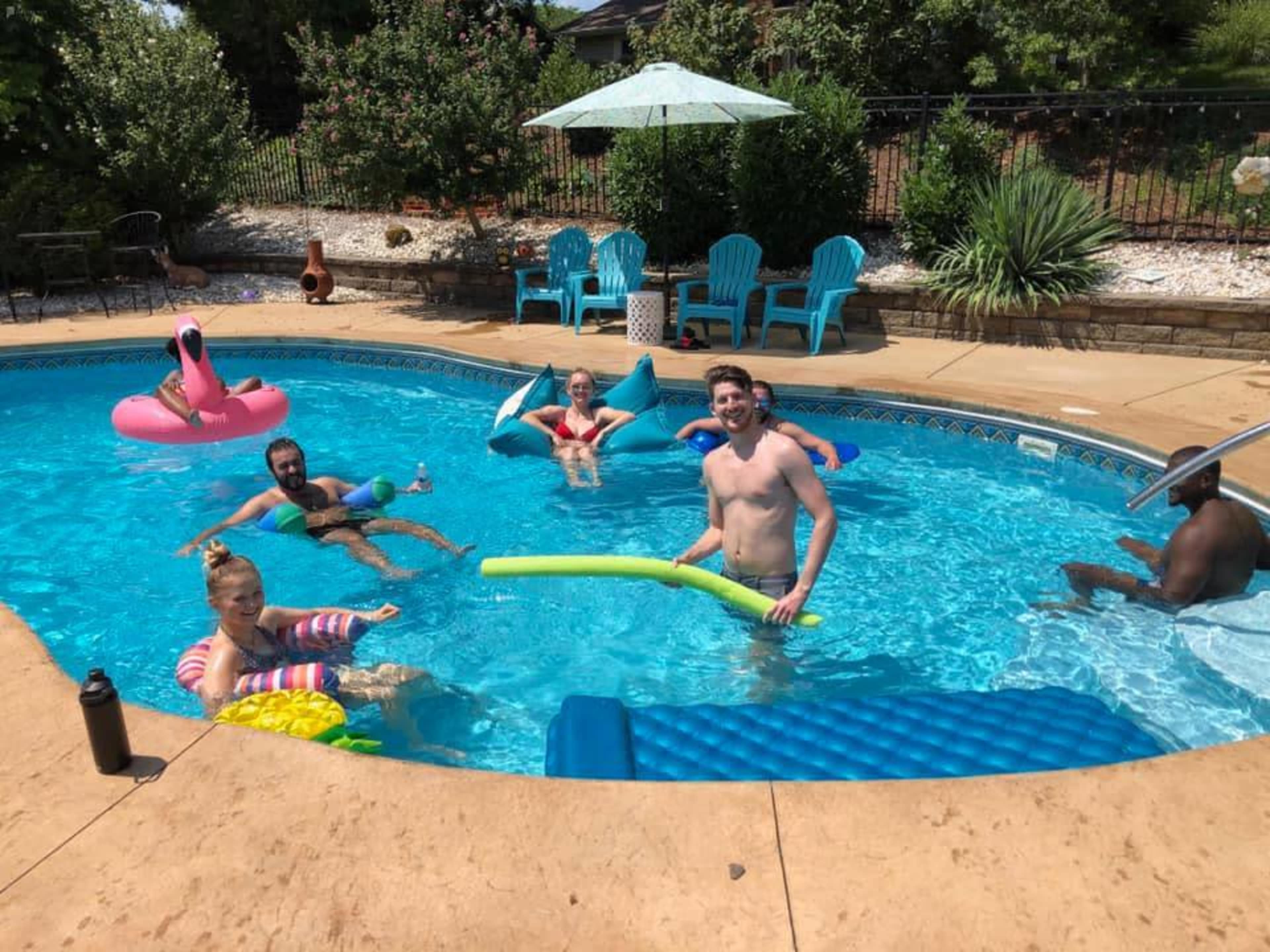 People are relaxing in a swimming pool surrounded by lounge chairs and a sun umbrella.
