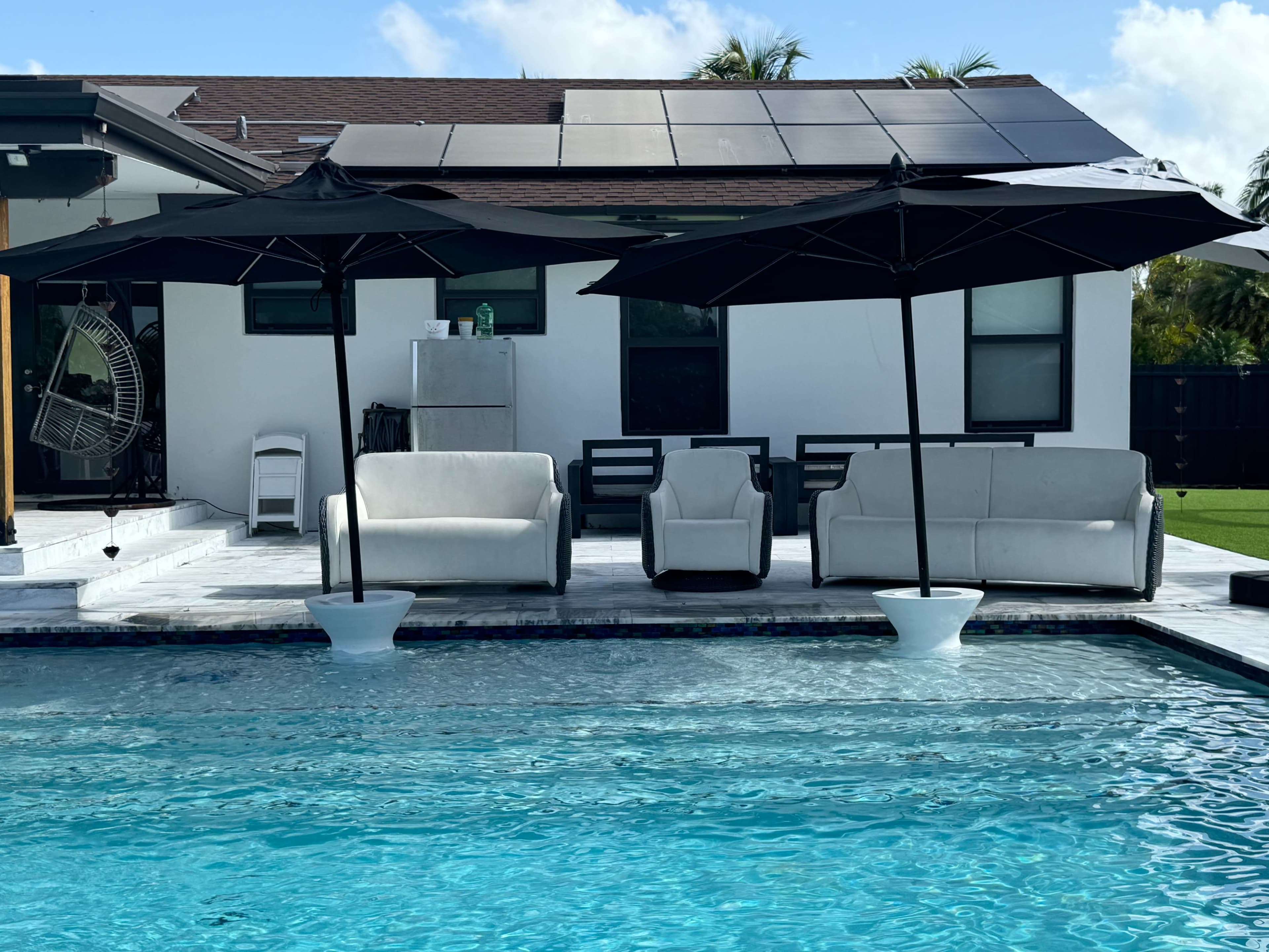 A swimming pool with two large black umbrellas over gray outdoor furniture next to a white modern house with solar panels on the roof.