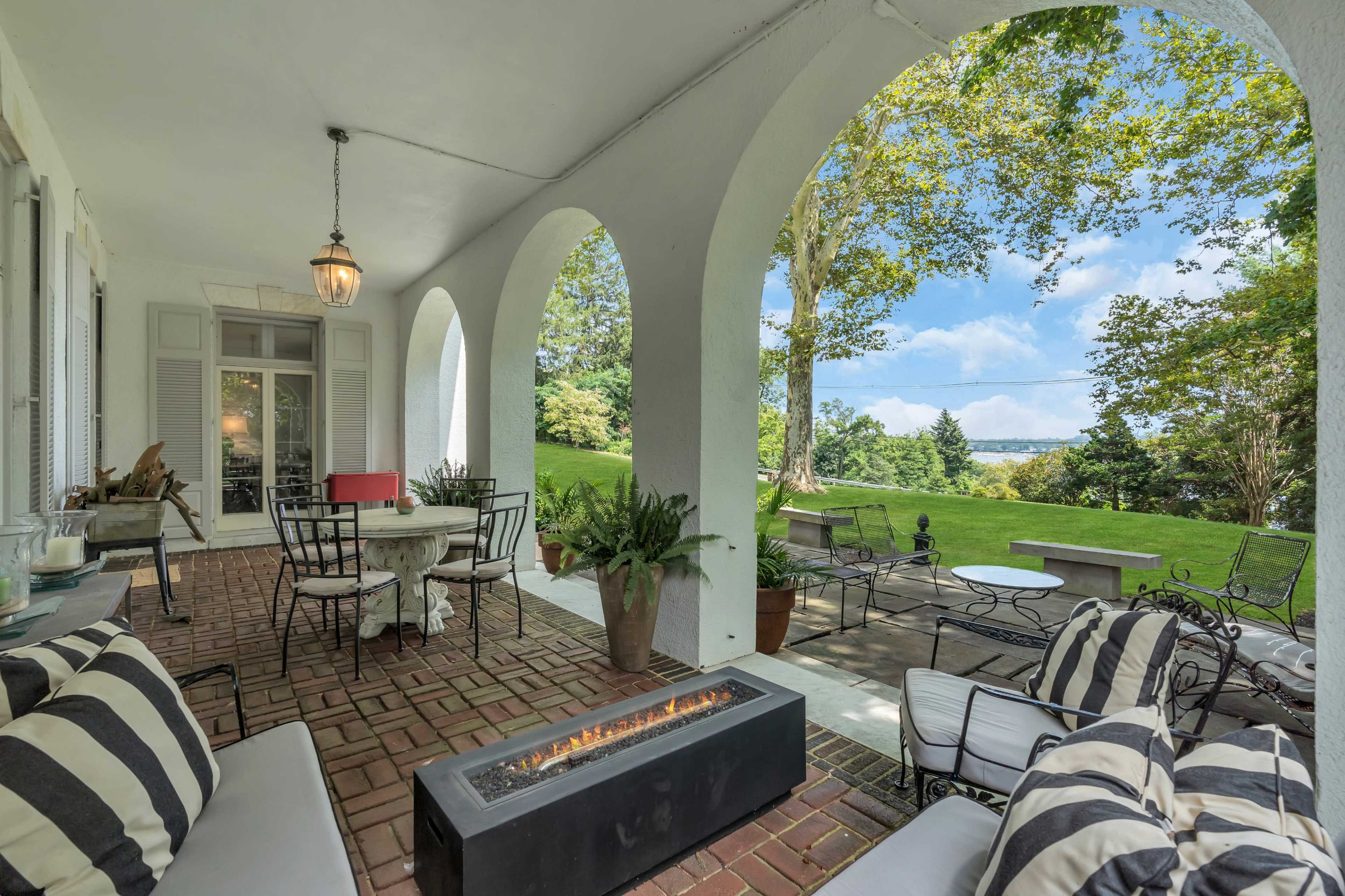 A covered patio features comfortable seating and a modern fire pit, framed by arched columns overlooking a green lawn and trees.
