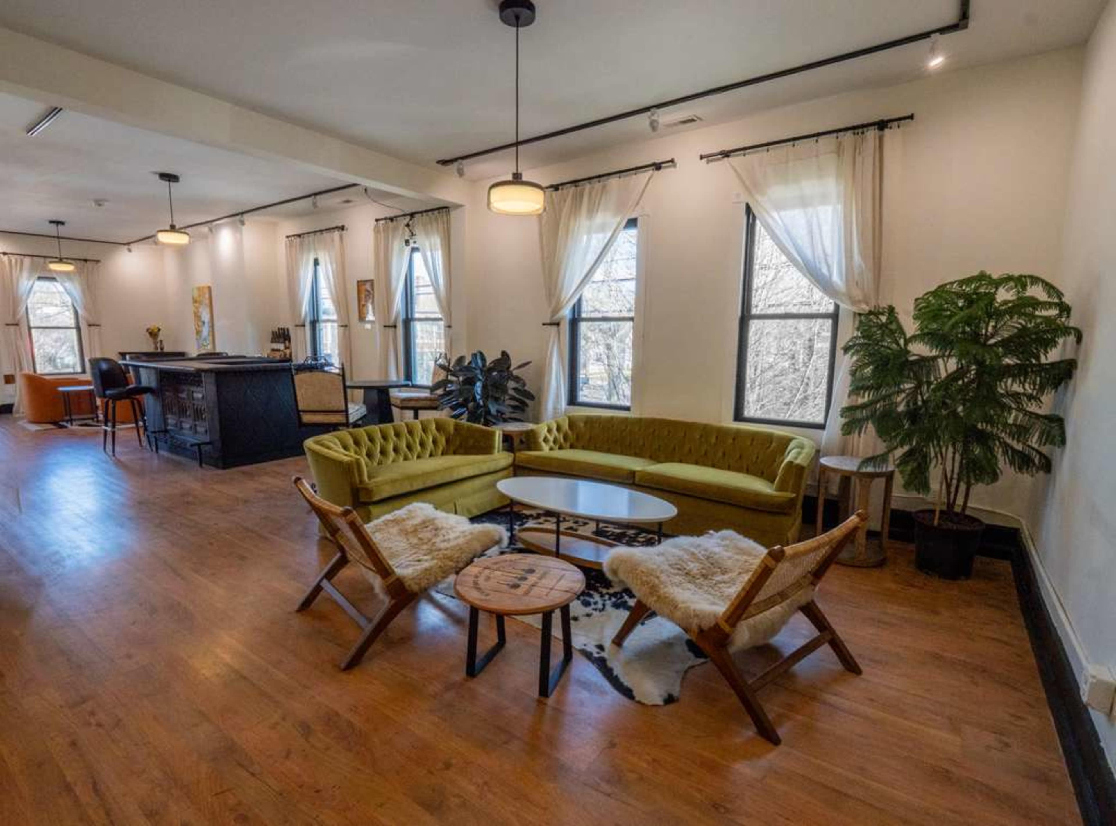 The image shows a spacious, well-lit living area featuring a yellow sofa, wooden flooring, and large windows, along with a small table surrounded by two chairs.