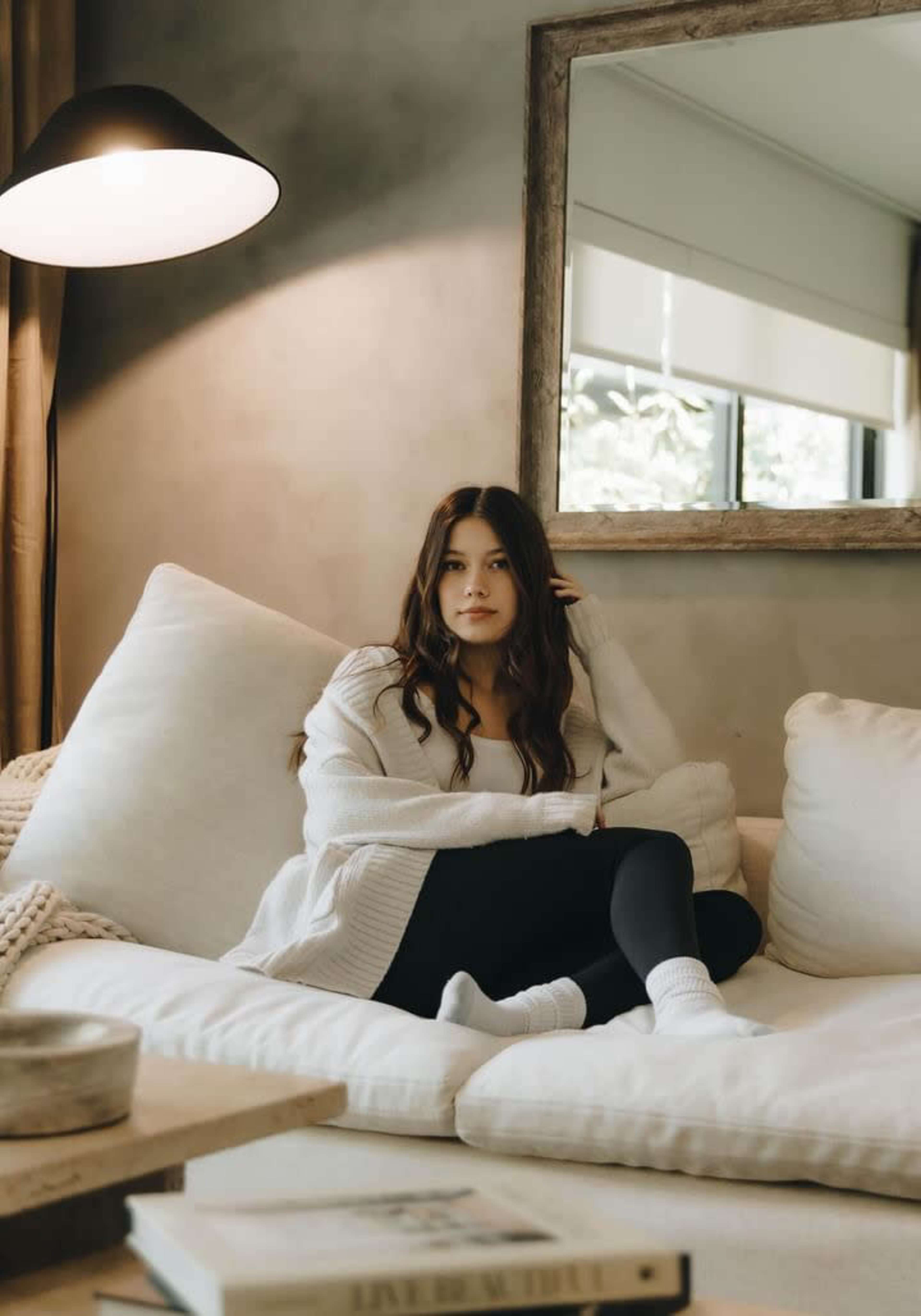 A woman sits on a large white sofa in a cozy living room, with a lamp and a mirror in the background.
