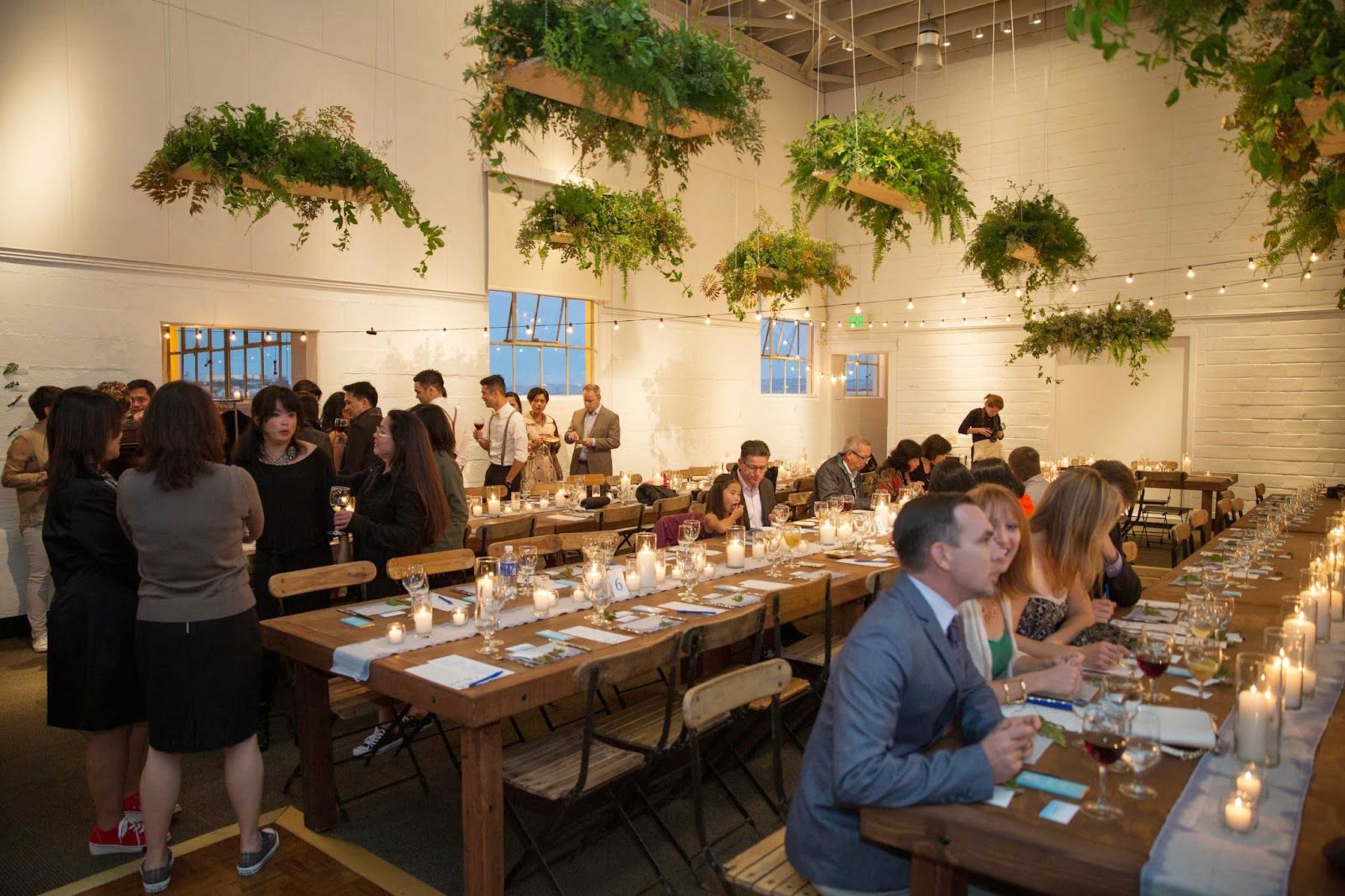 A gathering of people is seated at long tables decorated with candles and surrounded by hanging greenery in a well-lit venue.
