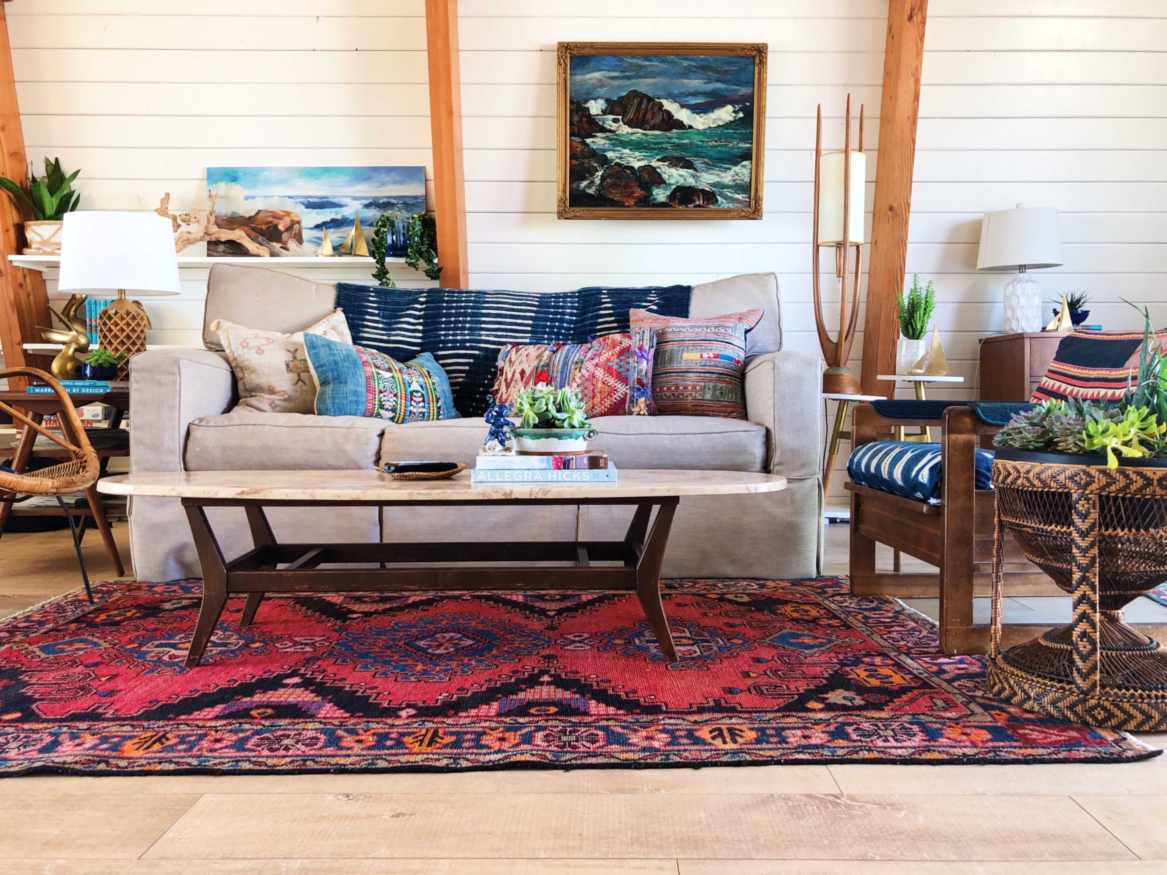 The image shows a living room with a beige sofa adorned with colorful pillows, a wooden coffee table on a patterned rug, and various decorative items on the surrounding furniture.