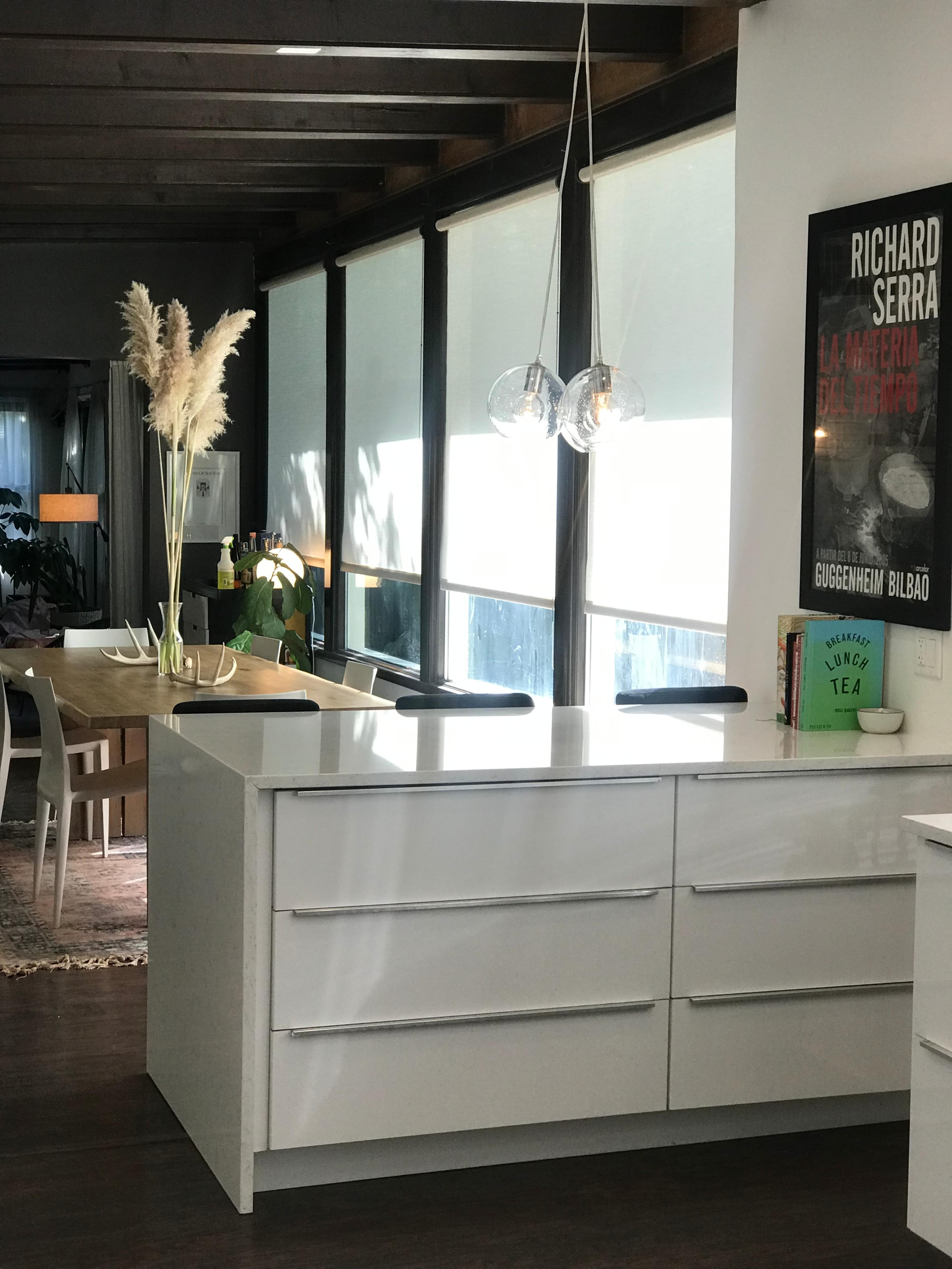 The image shows a modern kitchen with a white island and drawers, large windows allowing natural light, and a dining area in the background.