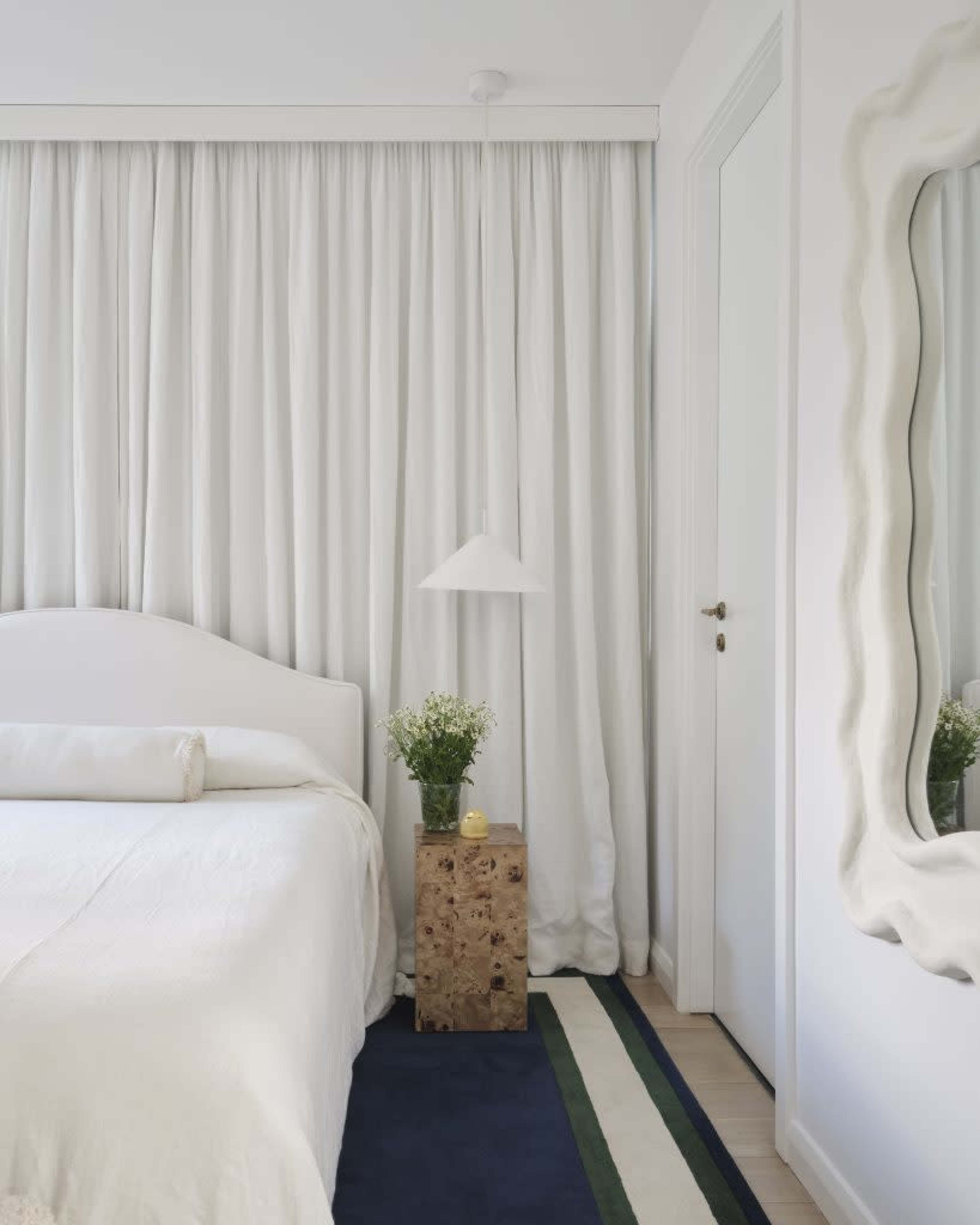 A bright bedroom features a white bed, a nightstand made of natural wood, and floor-to-ceiling white curtains.