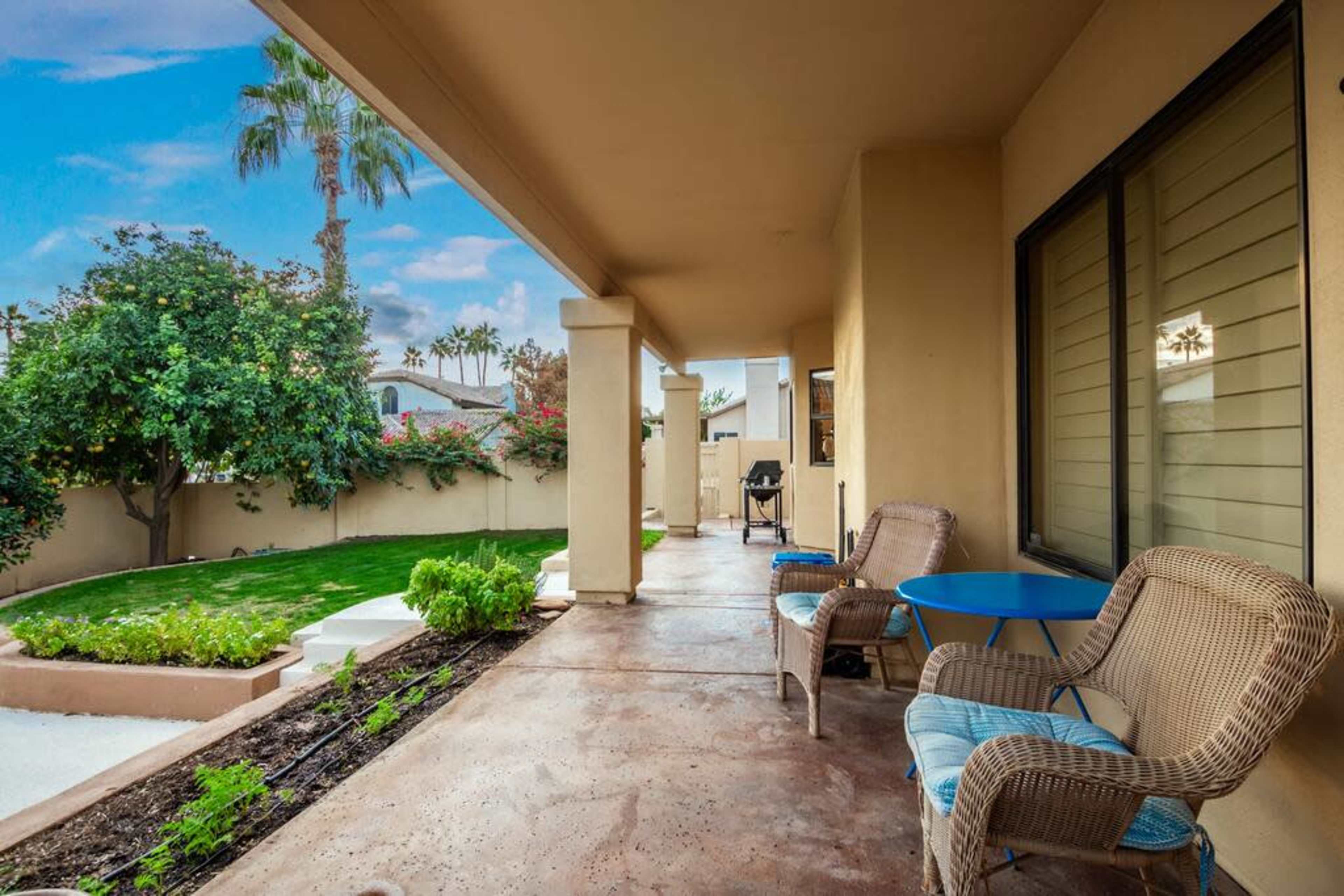 A covered patio features two wicker chairs and a small blue table, overlooking a green lawn and garden area.