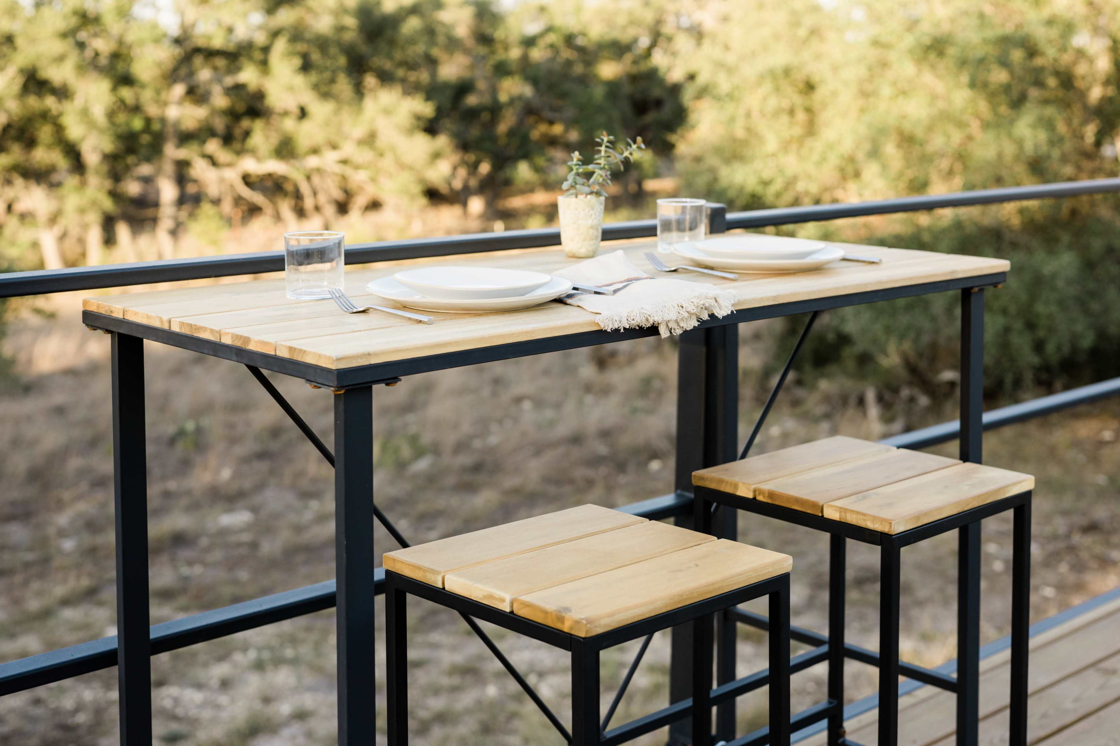 A wooden table with two stools is set for a meal on a balcony, surrounded by trees in the background.