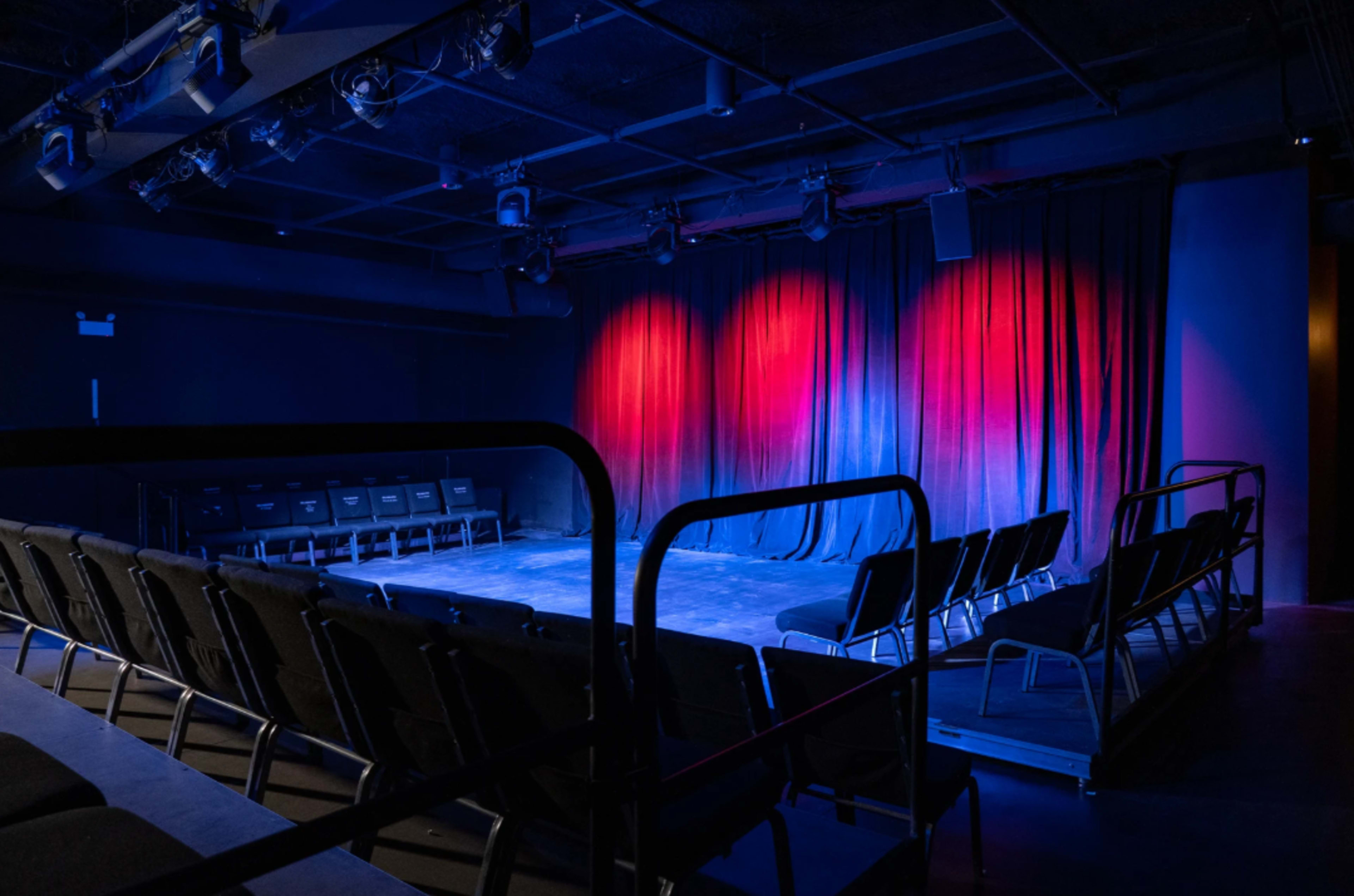 The image shows a dimly lit theater with a stage draped in red and blue curtains, surrounded by rows of black seating.