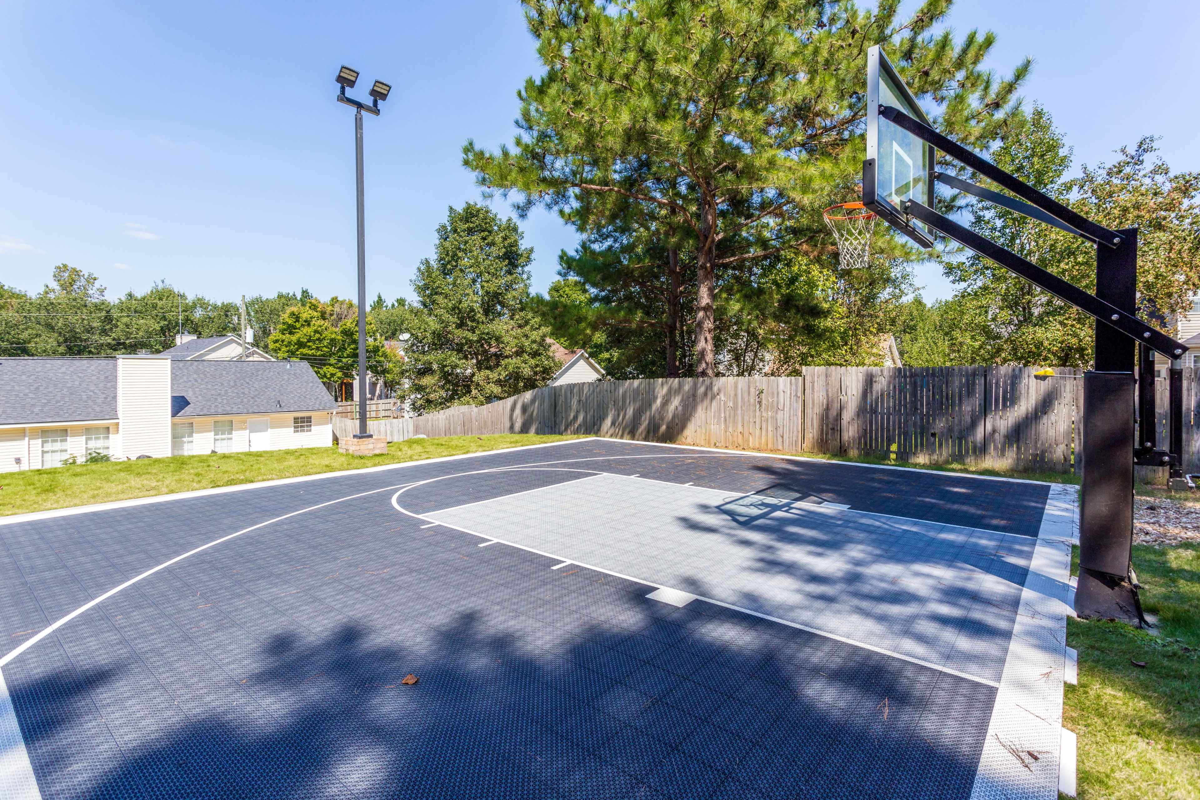 An outdoor basketball court surrounded by a wooden fence and trees, with a basketball hoop mounted on a black support structure.