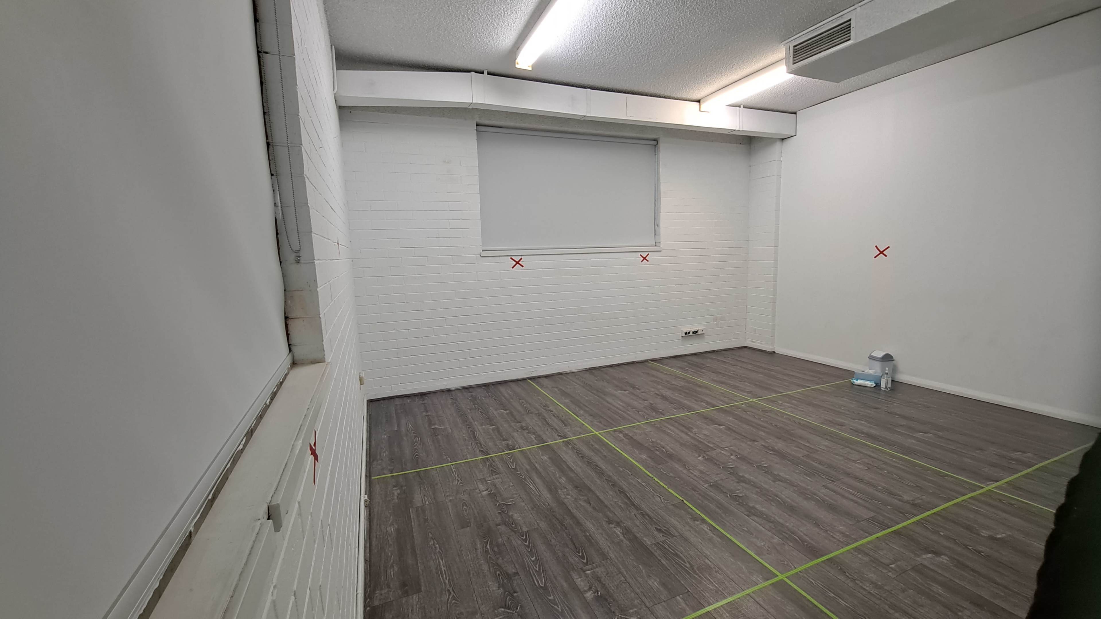 An empty room with white brick walls, a window covered by a blind, and marked floors in green tape with red 'X' indicators.
