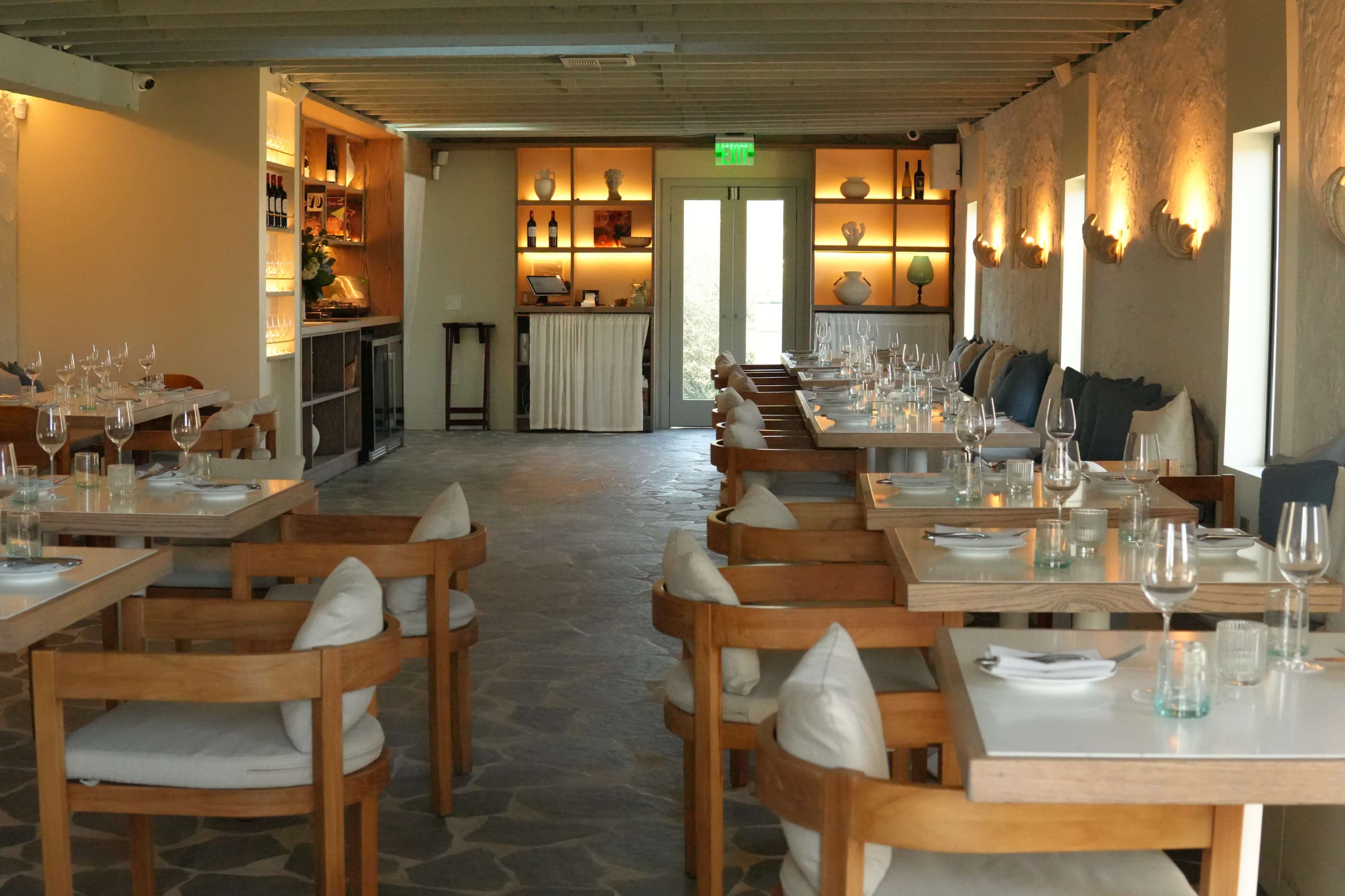 The image shows an empty restaurant interior with wooden tables set for dining and soft lighting illuminating the space.