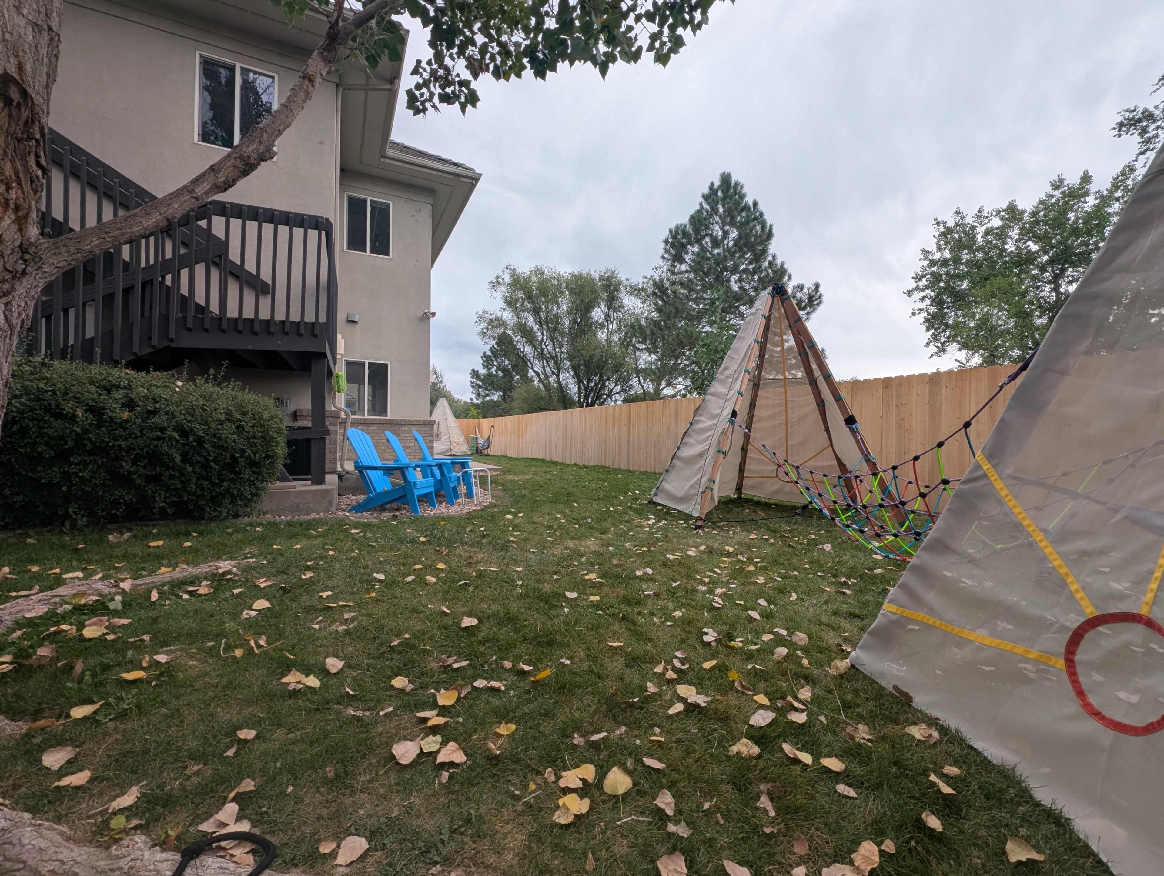 The image shows a backyard with a grassy area, a tree, a wooden fence, and two blue chairs facing a colorful play structure resembling a tent or teepee.