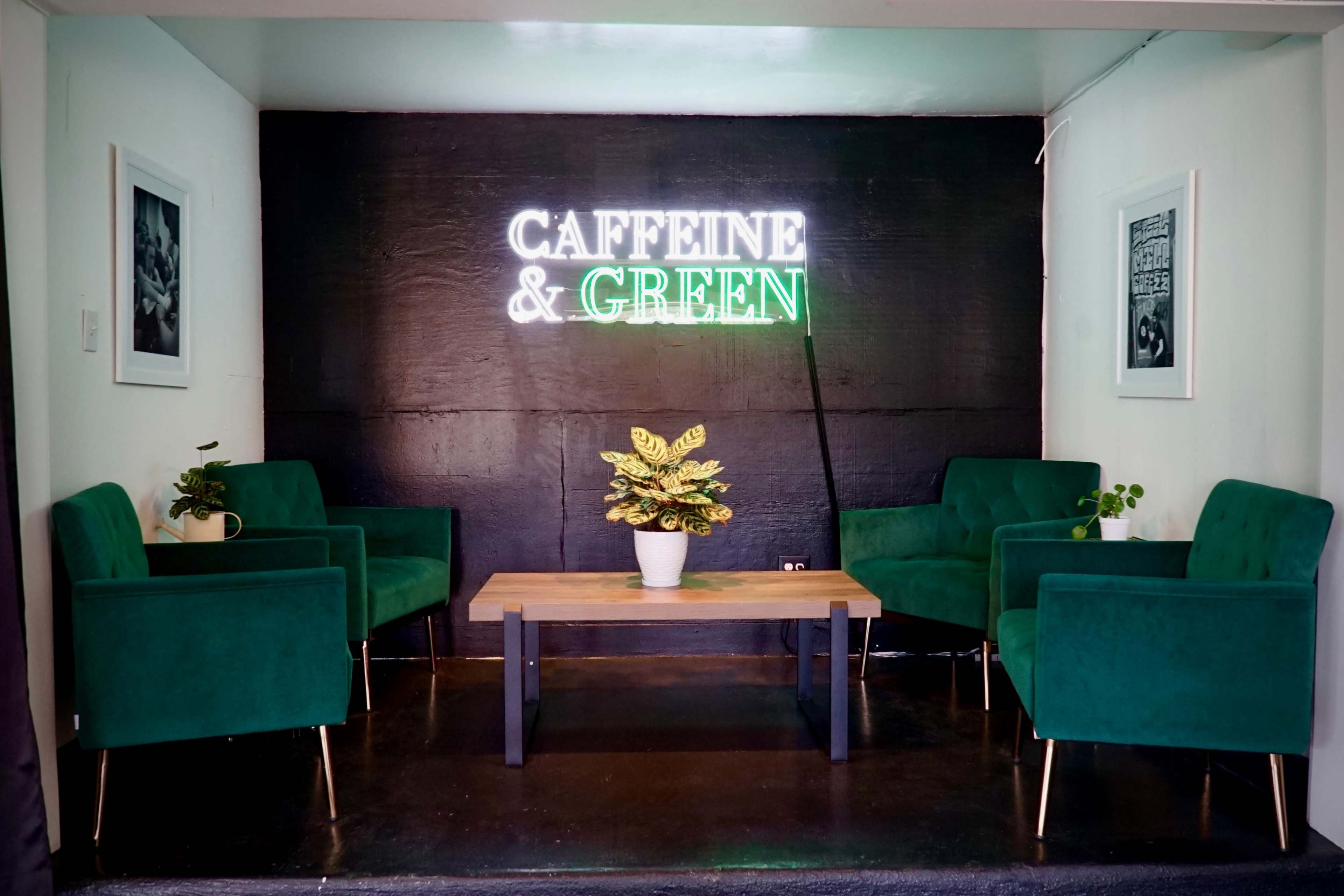 A seating area with four green chairs and a wooden table, illuminated by a neon sign that reads "CAFFEINE & GREEN."