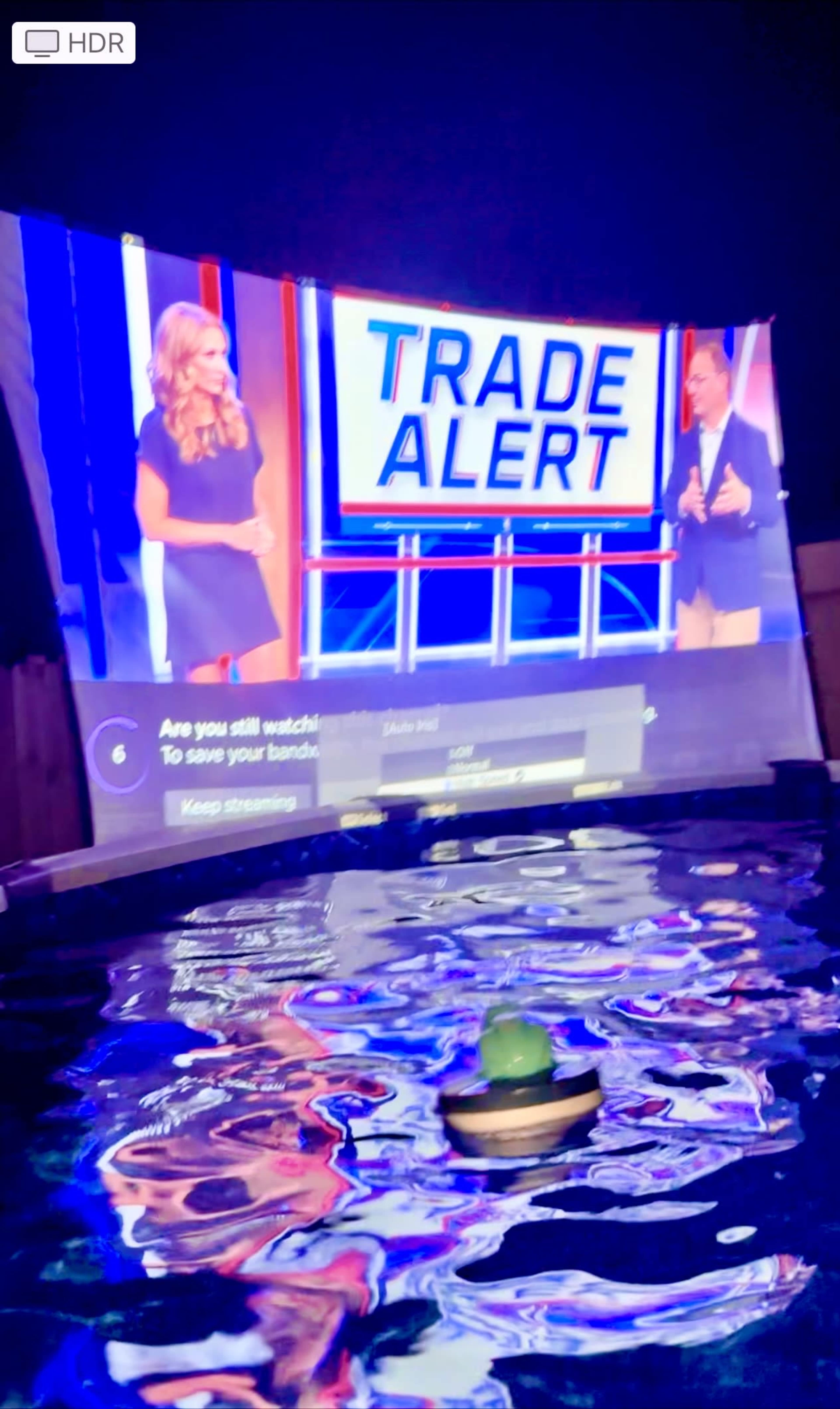 A large screen displays a "Trade Alert" message next to two presenters, reflecting in the water of a pool at night.