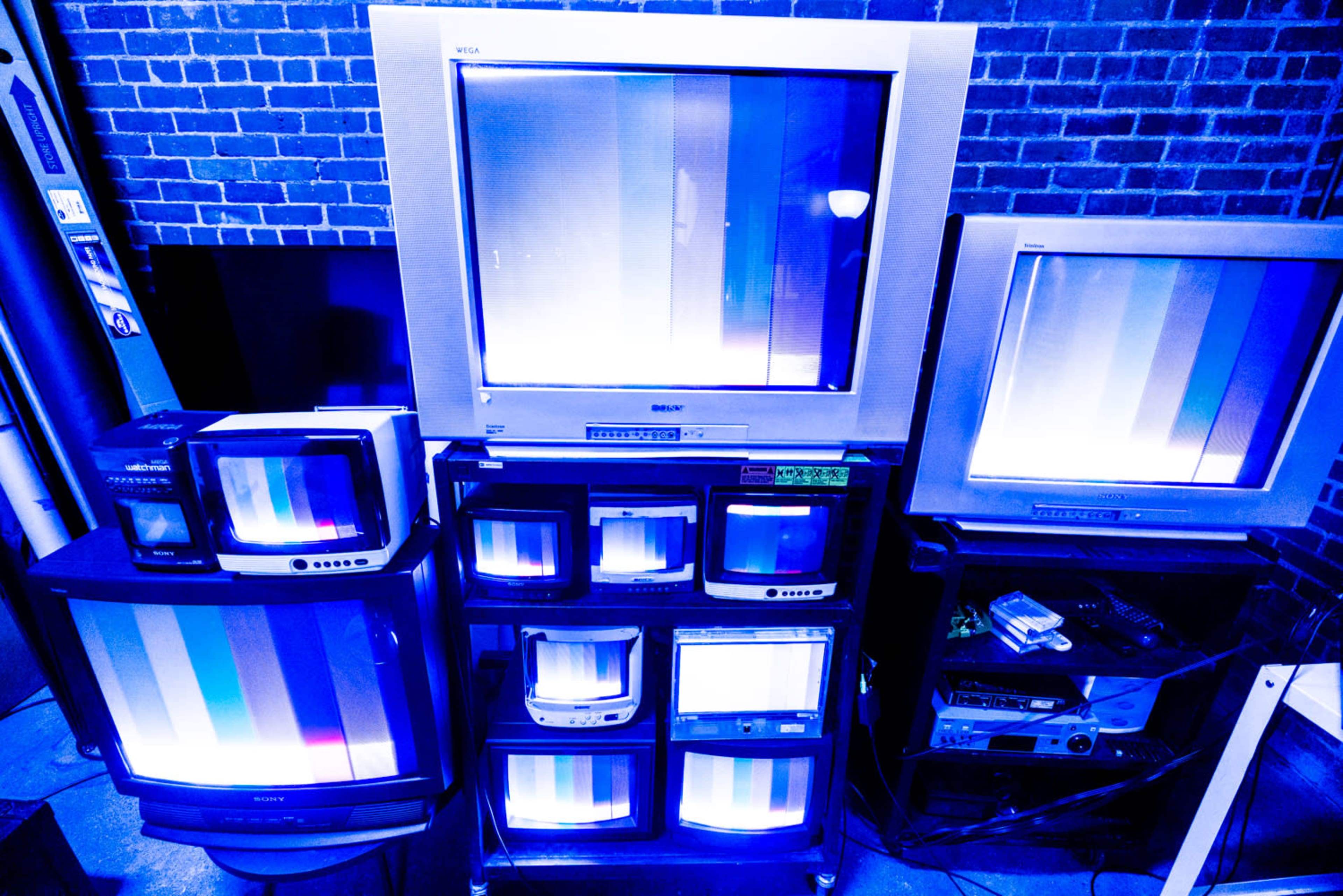 A collection of vintage televisions displaying static and bright vertical lines is arranged against a brick wall.