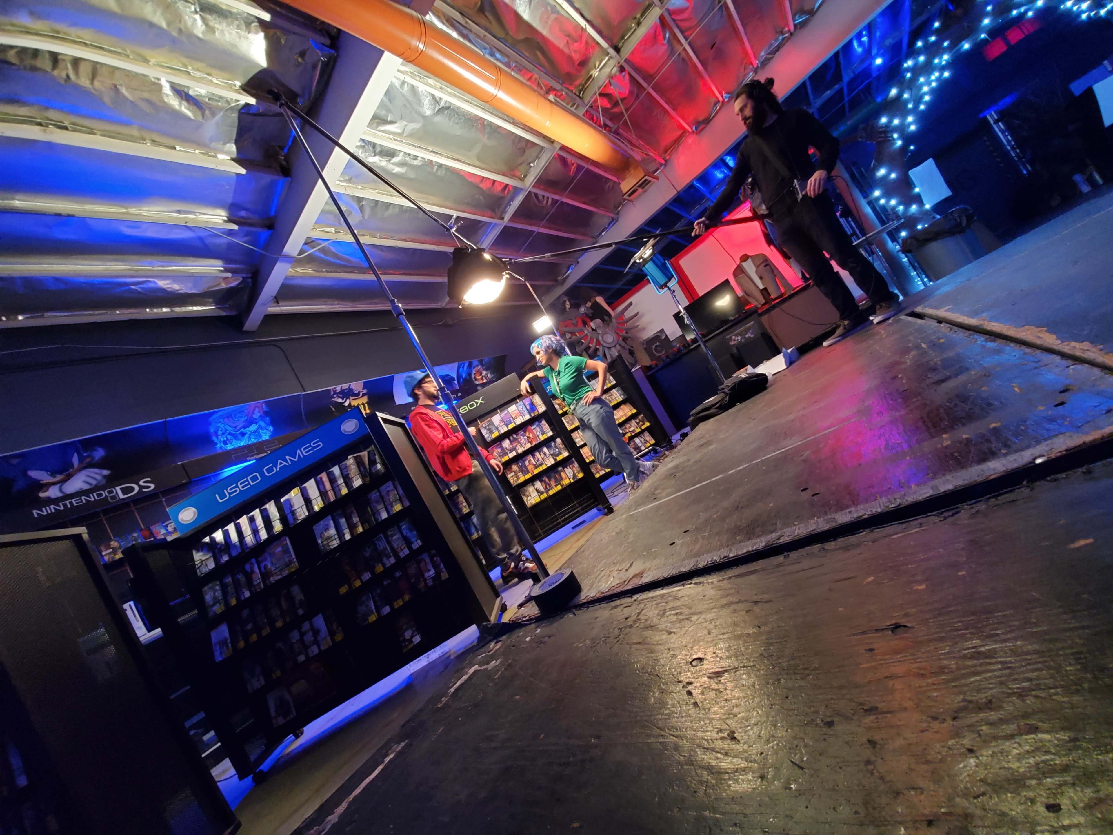 A brightly lit gaming store interior with two people standing near shelves filled with video game cases, while a third person adjusts equipment on the floor.
