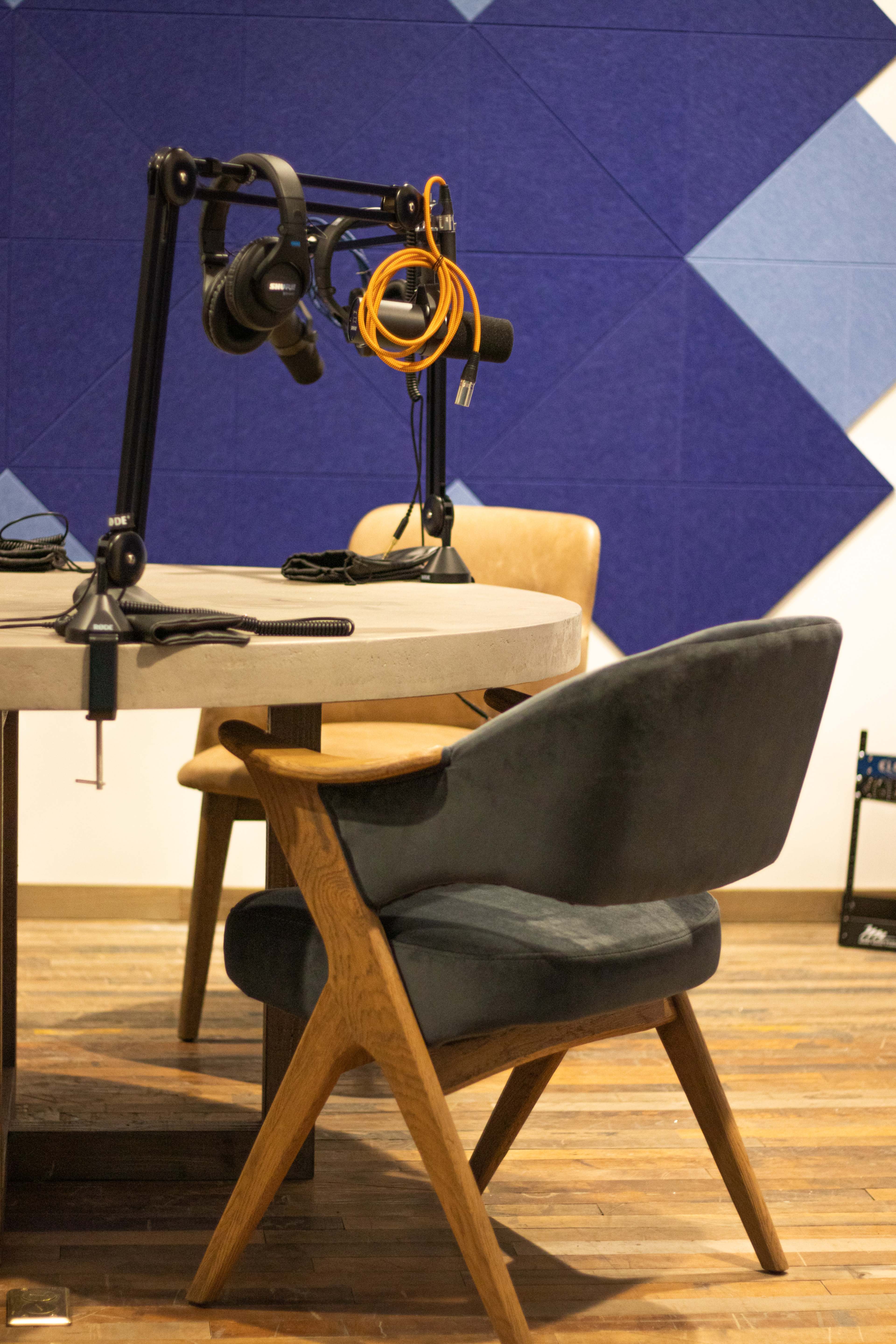 The image shows a recording studio setup with a round table, two chairs, and a microphone arm with headphones hanging from it against a patterned blue wall.