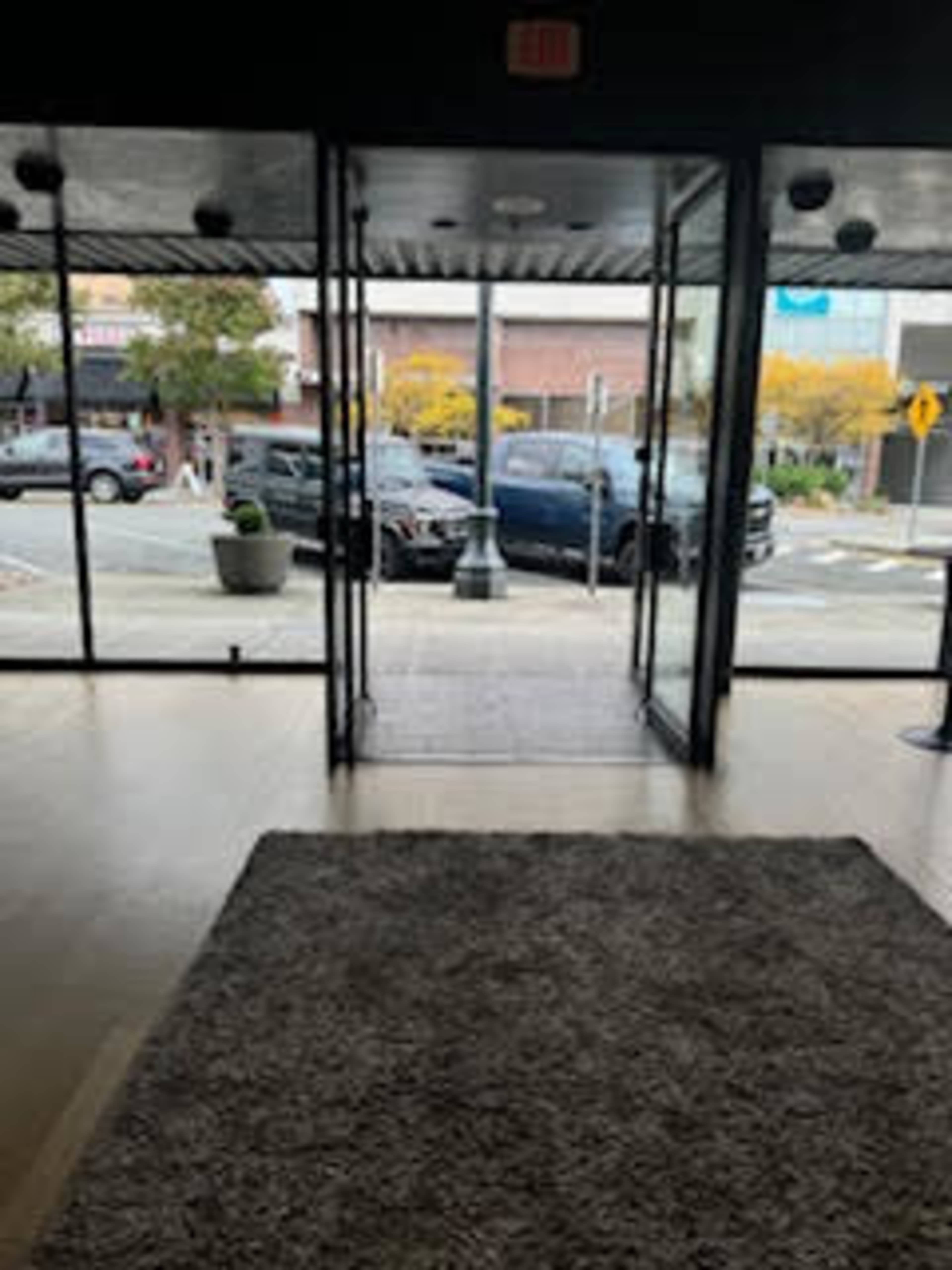 The image shows the interior of a building with glass doors, leading out to a street lined with parked cars and some greenery.