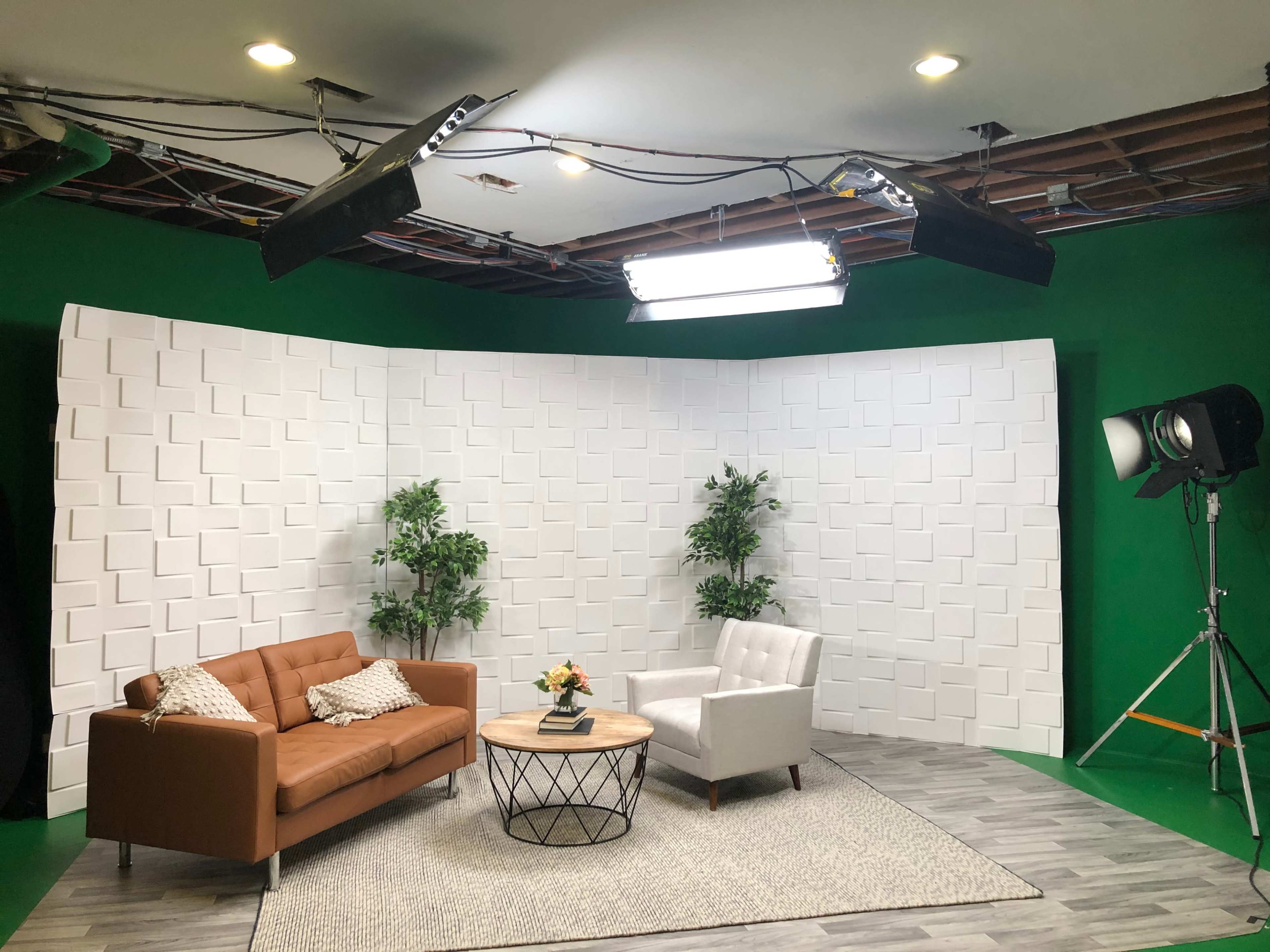 A neatly arranged studio setting with a brown sofa, a white armchair, a round coffee table with a flower vase, and a green screen backdrop.