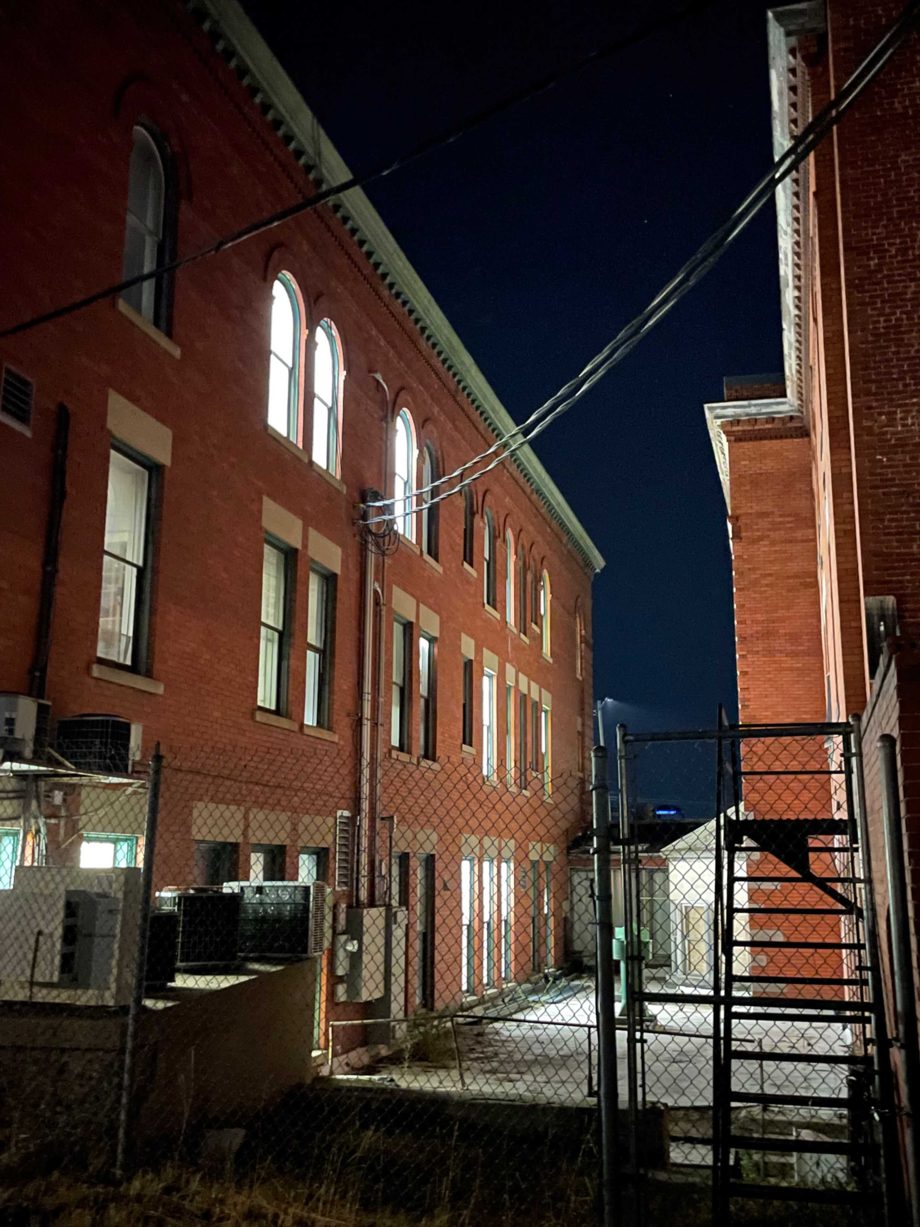 The image shows a narrow alleyway between two red brick buildings at night, illuminated by the windows and streetlights.