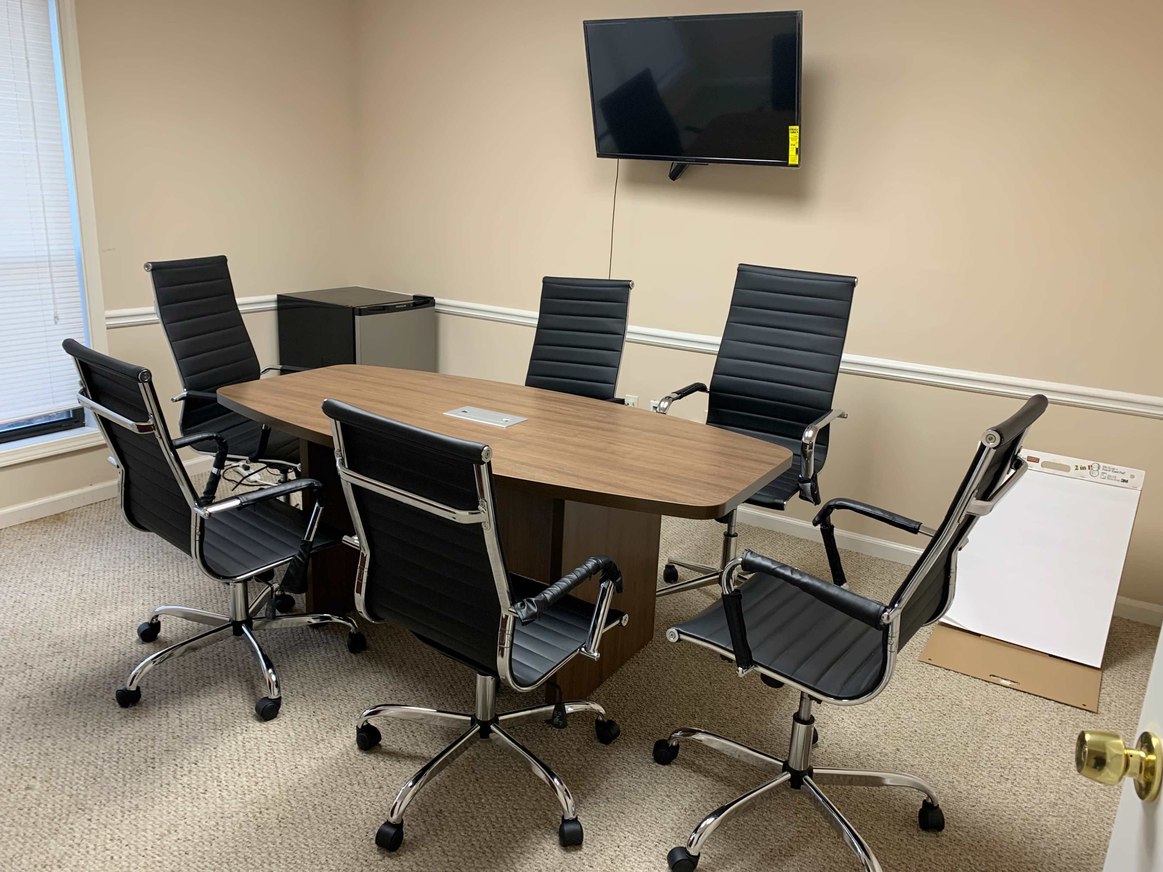 A conference room is furnished with a rectangular wooden table surrounded by six black rolling chairs and features a wall-mounted television.