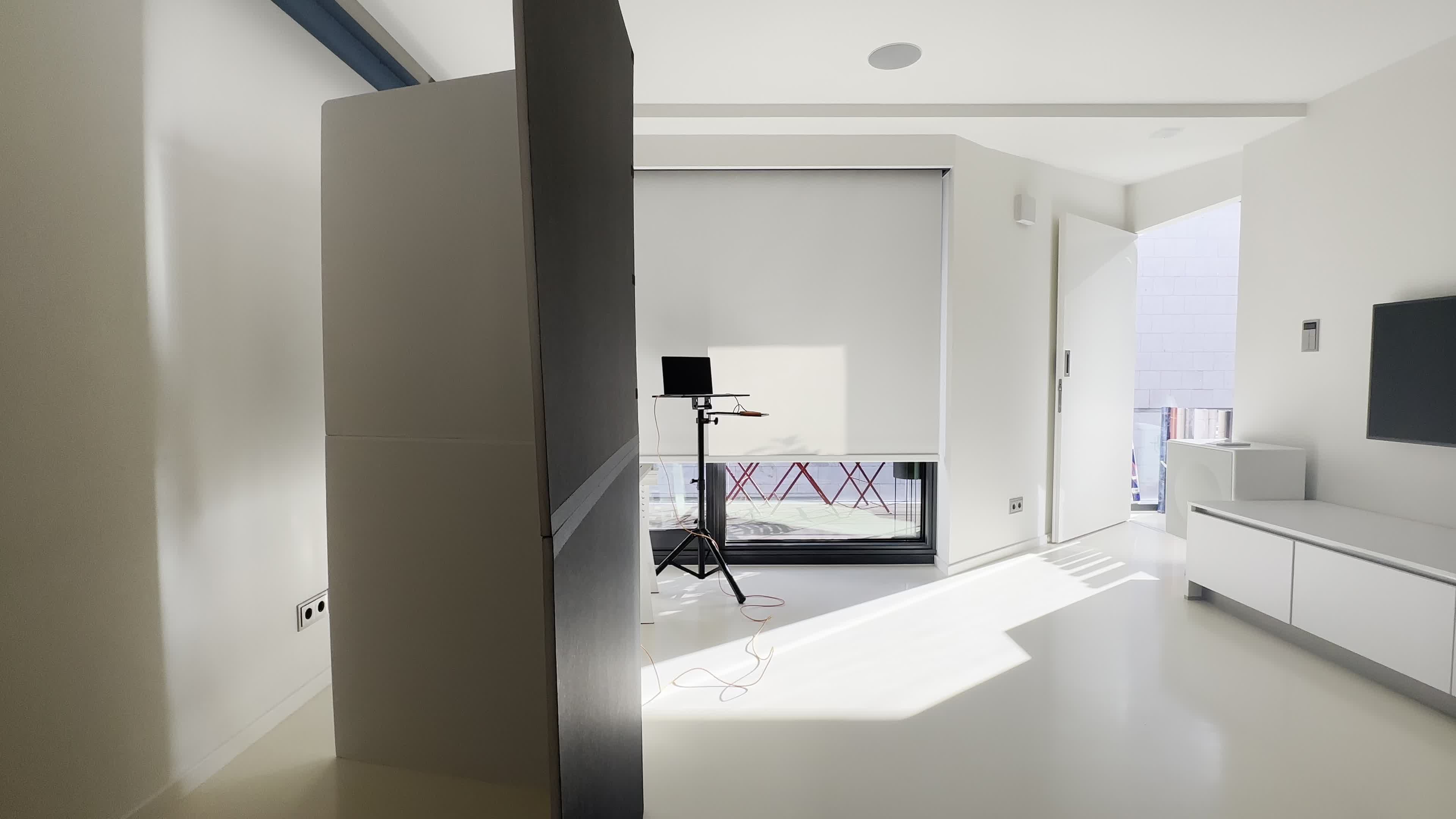 A modern, minimalistic interior space with a partition on the left, a laptop on a stand, and large windows allowing natural light to enter.