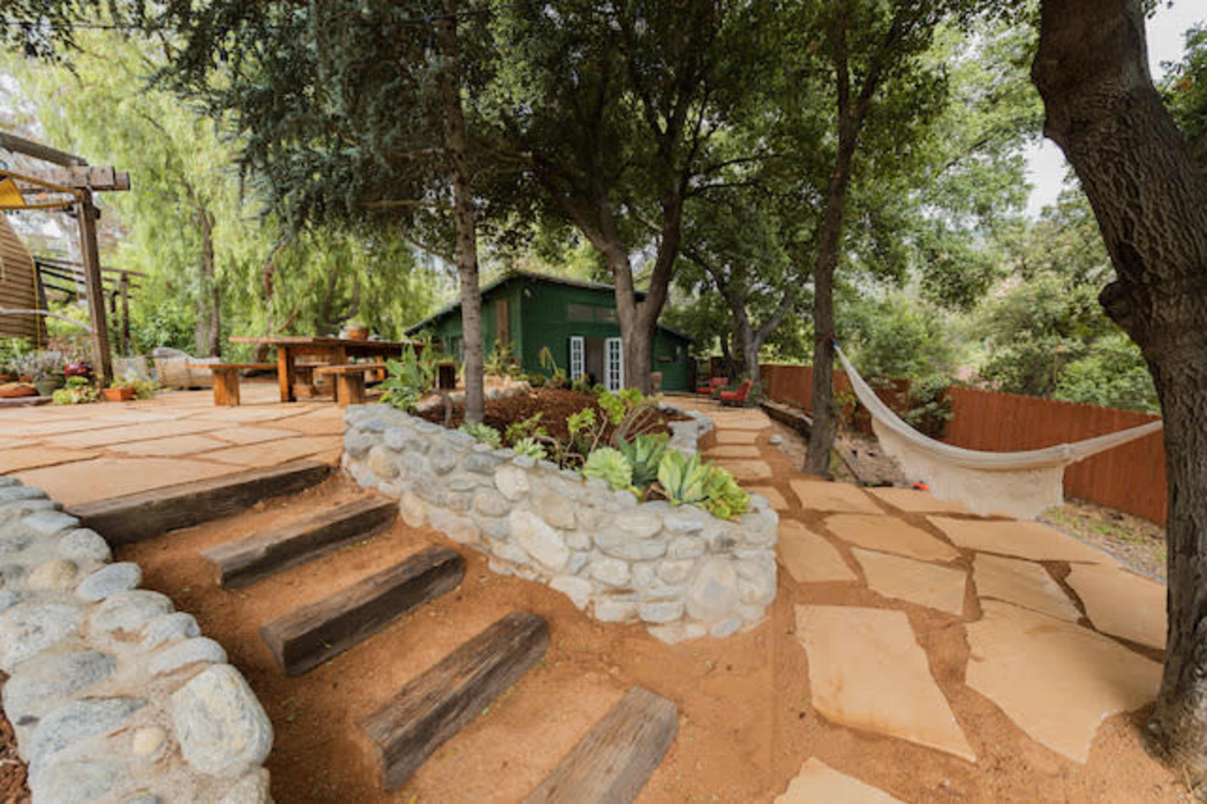 The image shows a landscaped garden with stone paths, a wooden table, and a hammock nestled between trees.