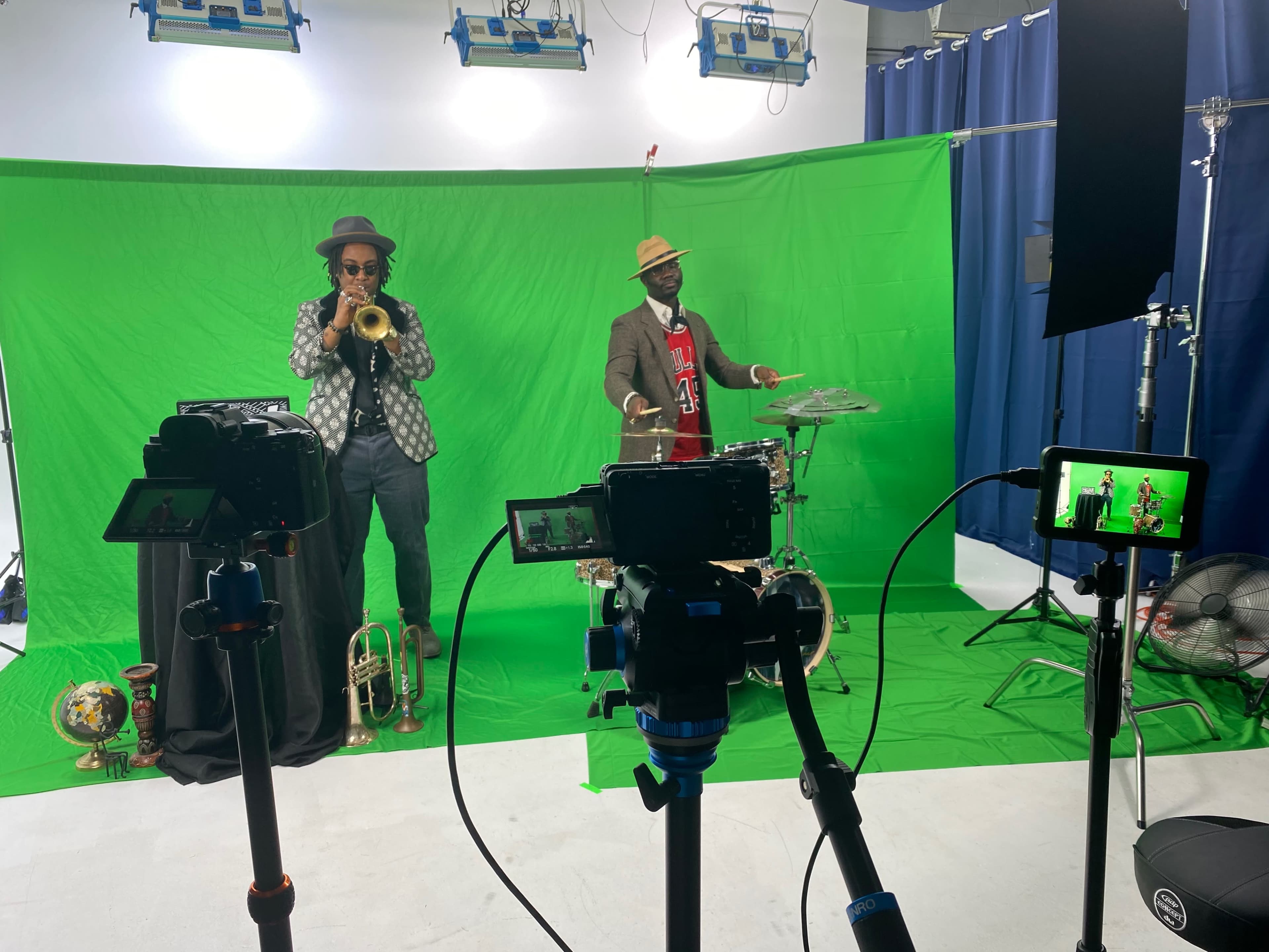 Two musicians perform in front of a green screen, one playing the trumpet and the other playing the drums, while several cameras are positioned in the foreground.