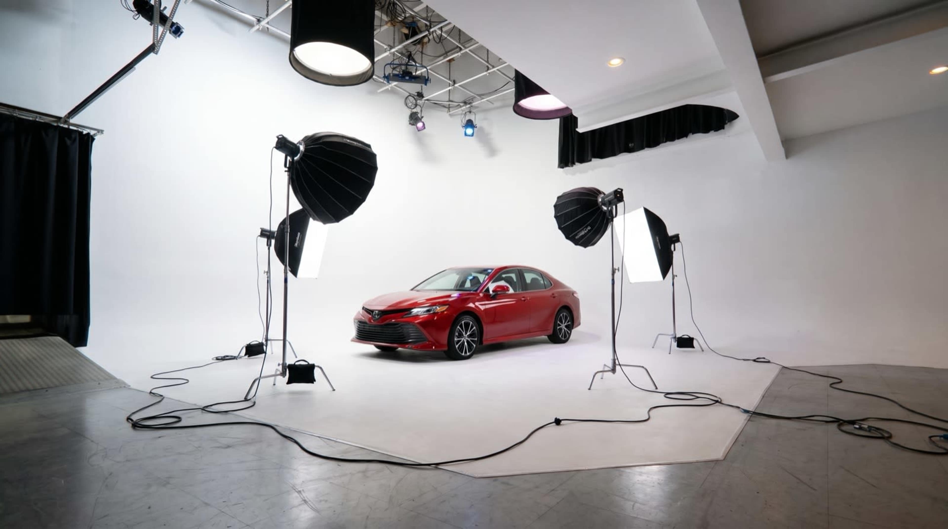 A red car is displayed in a photo studio surrounded by large lighting equipment and a plain white backdrop.