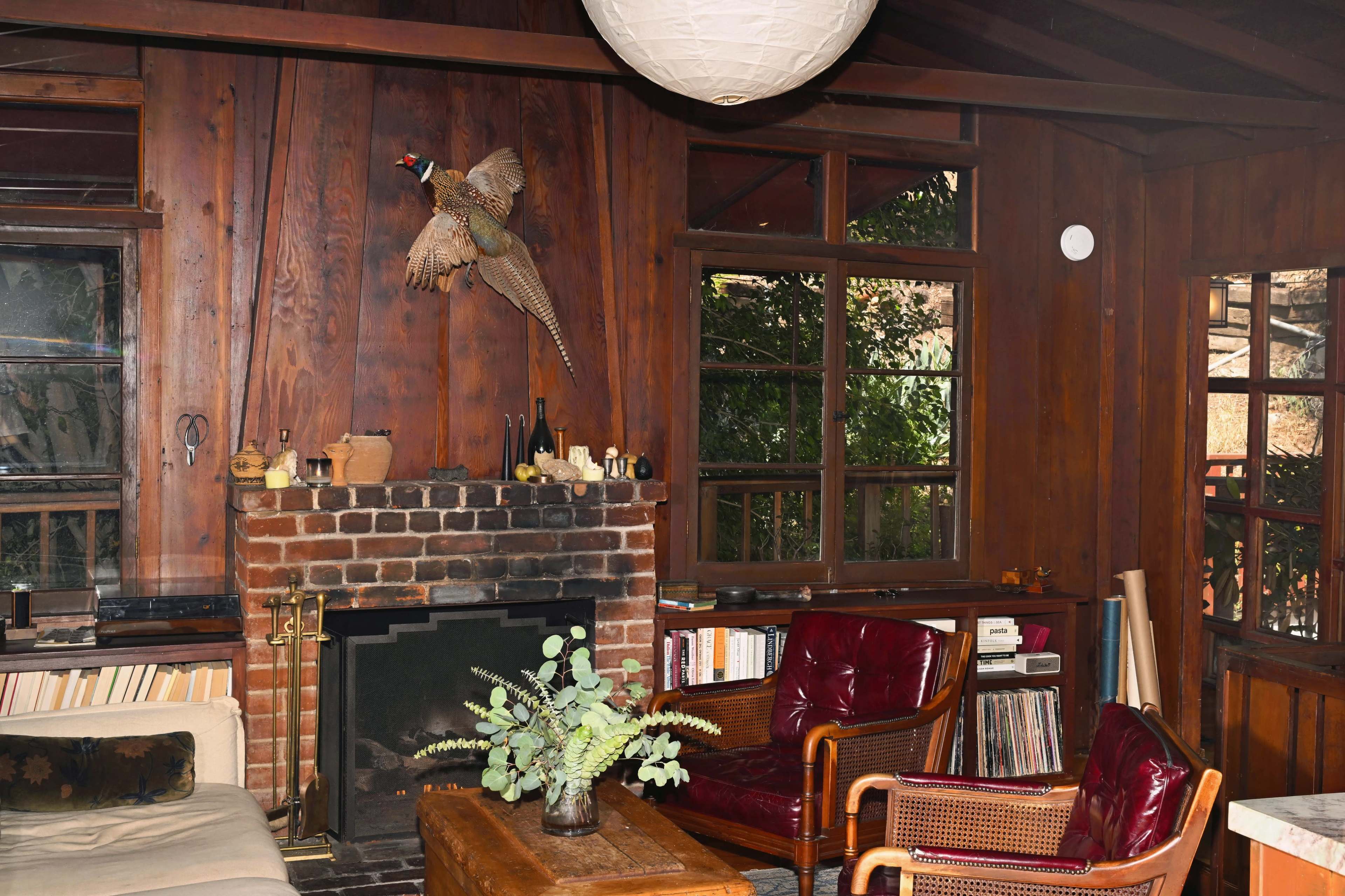 The image shows a cozy wooden interior of a room featuring a brick fireplace, a mounted bird on the wall, and comfortable seating arrangements with a coffee table.
