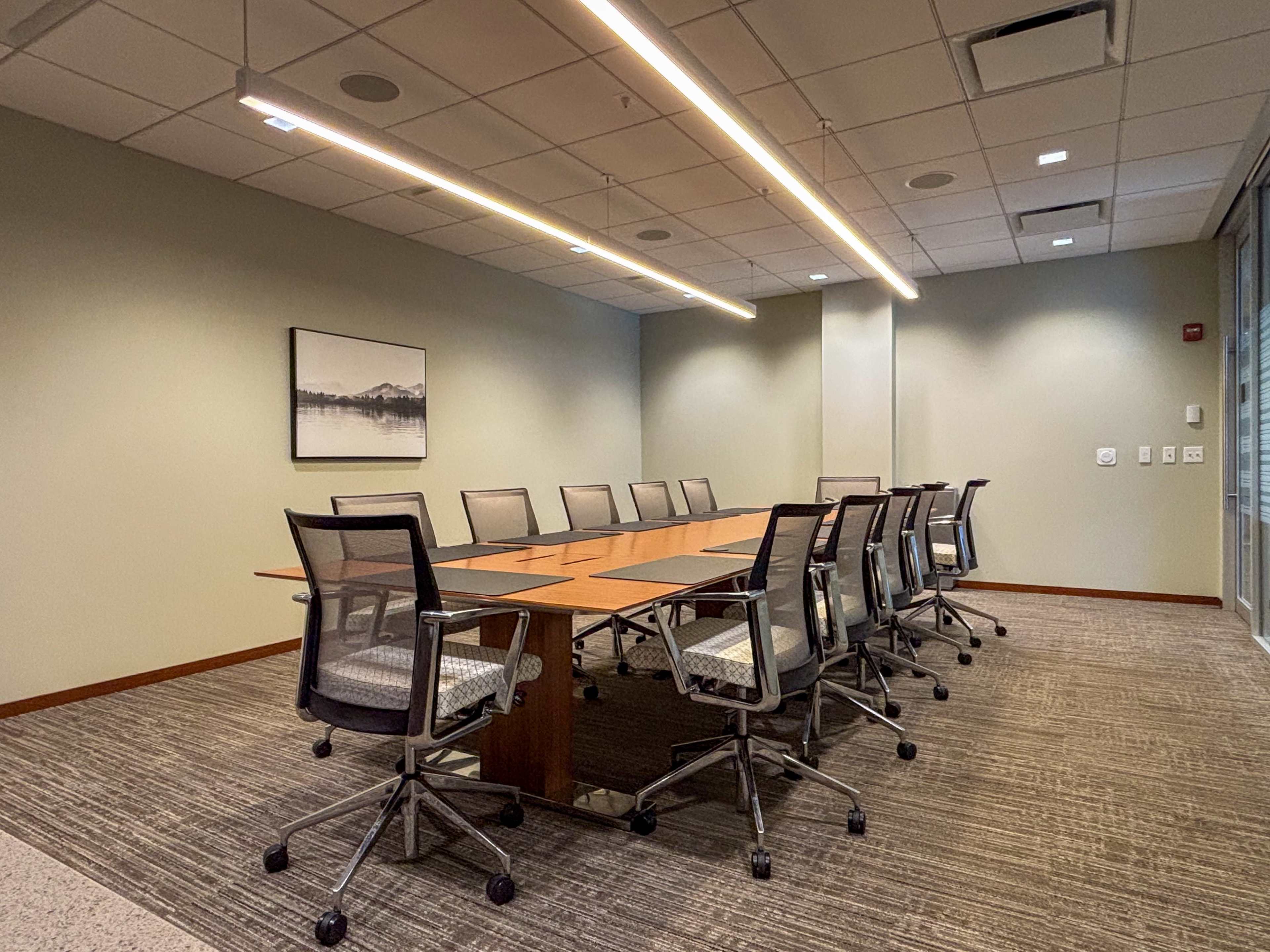 The image shows a modern conference room with a long table surrounded by ergonomic chairs, illuminated by overhead lighting.