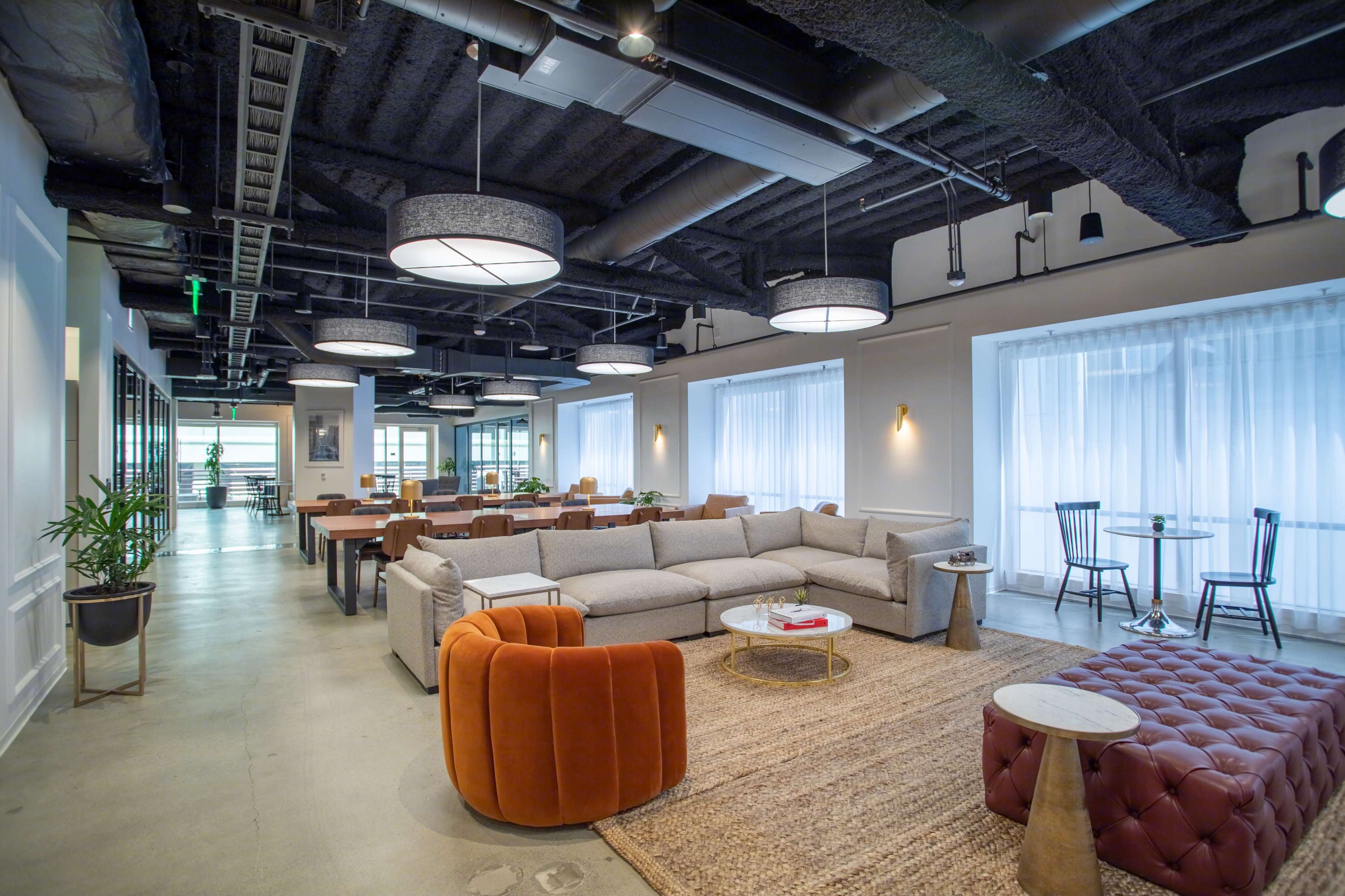 A modern lounge area featuring a gray sectional sofa, orange accent chairs, and a variety of tables under circular ceiling lights.