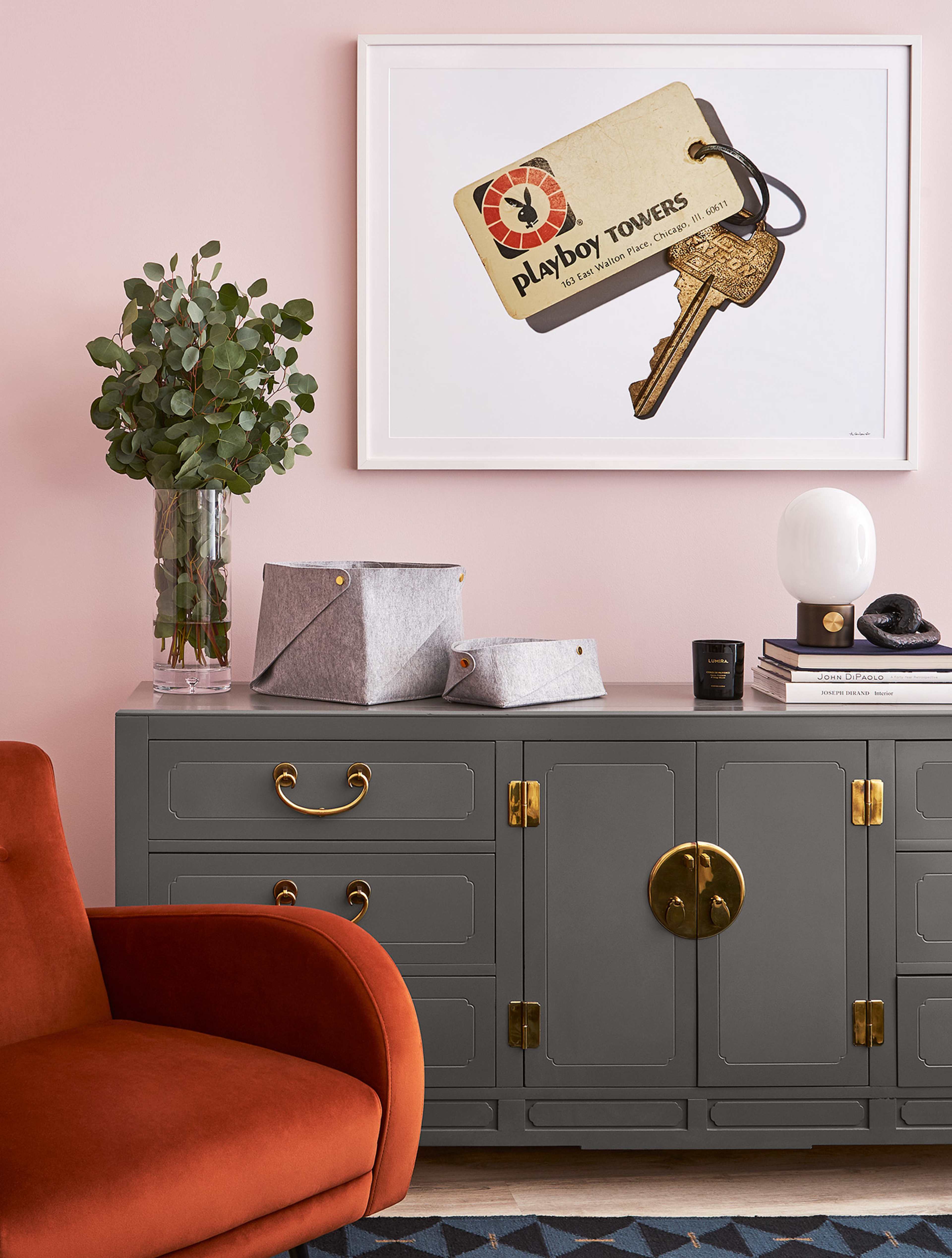 A stylish living room features a gray cabinet with gold hardware, a large framed art piece above it, a glass vase with eucalyptus, and a red armchair.